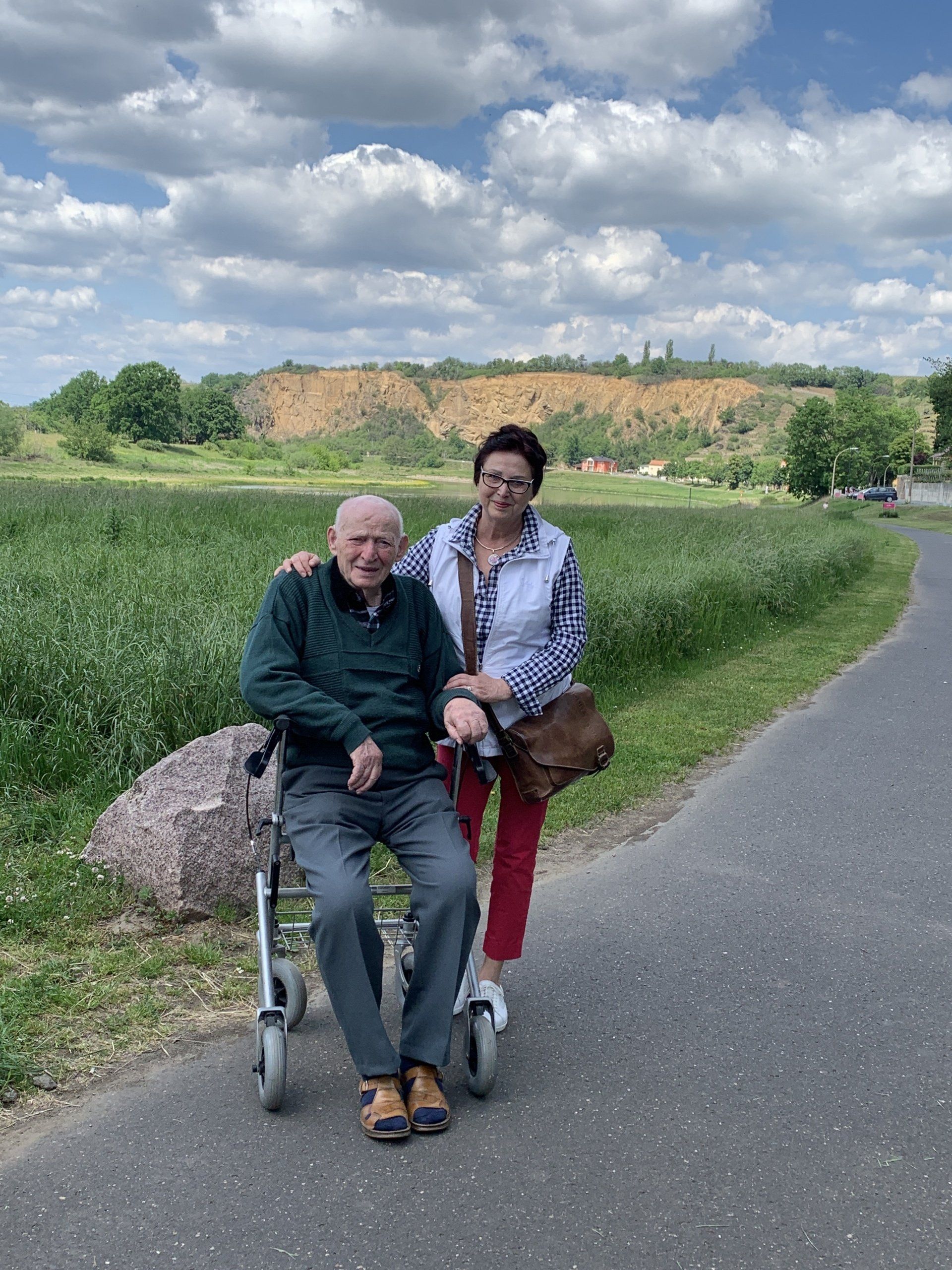 Eine ältere Person sitzt in einem Rollator, neben ihr eine Frau, auf einem Weg in einer parkähnlichen Umgebung.