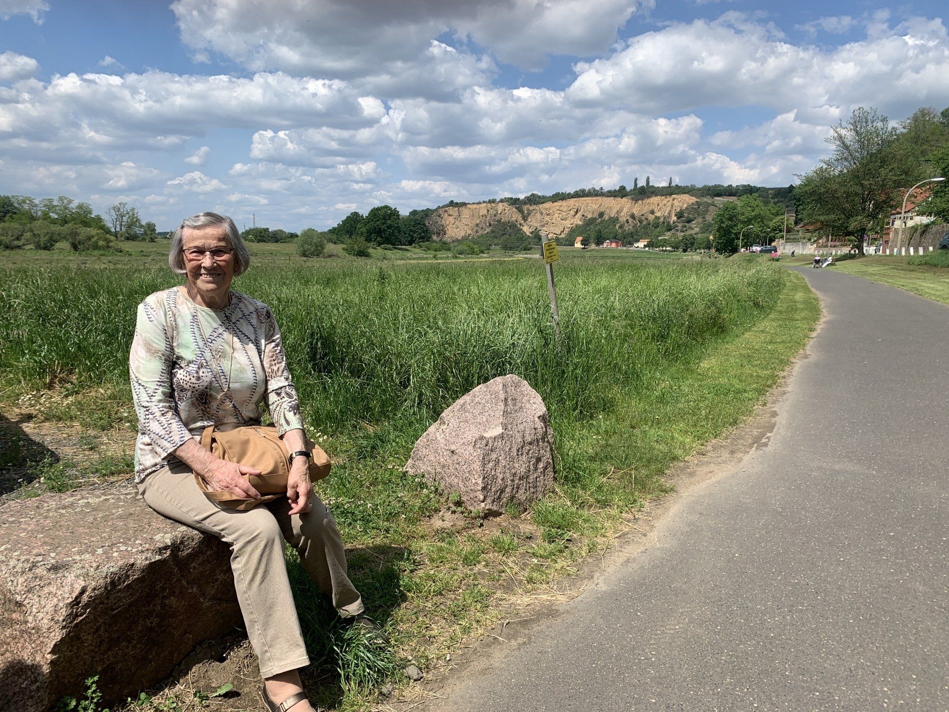 Frau sitzt auf einem Felsen an einem Pfad neben einem Feld. Sonniger Tag, blauer Himmel mit Wolken.