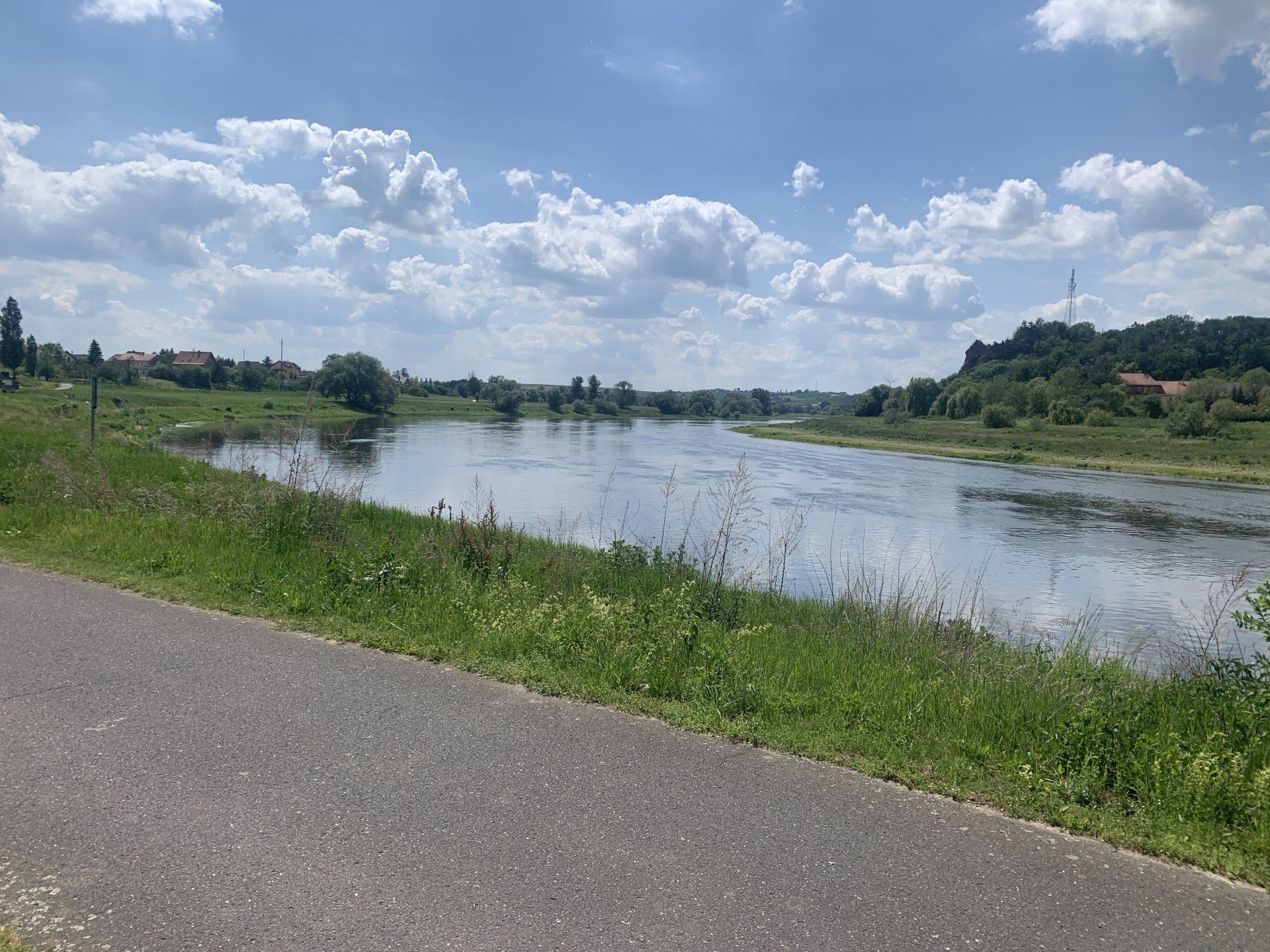 Flussszene unter teilweise bewölktem Himmel. Asphaltweg im Vordergrund, grünes Gras und Bäume am Wasserrand.