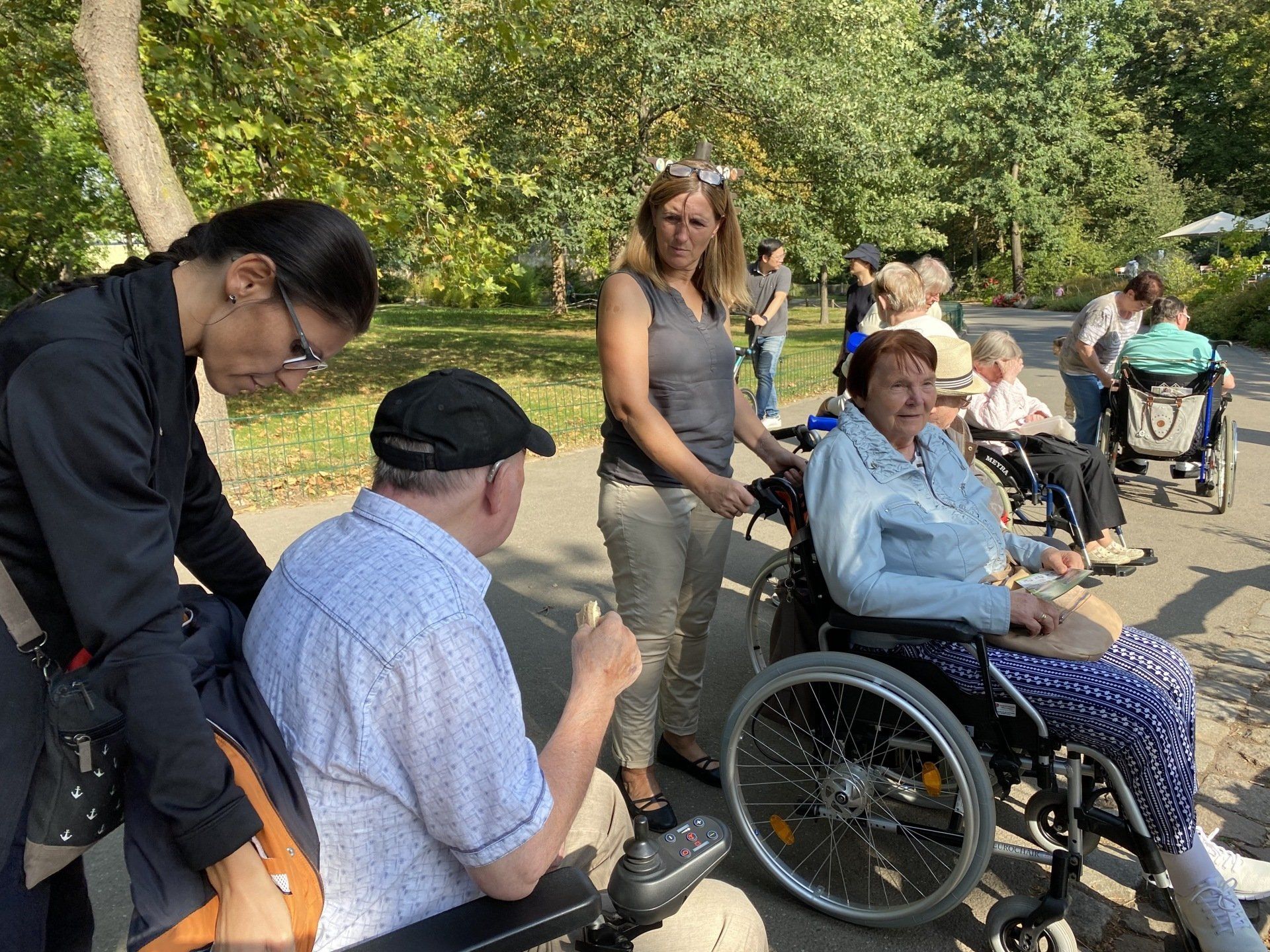 Menschen im Rollstuhl mit Betreuern im Freien; sonniger Tag.