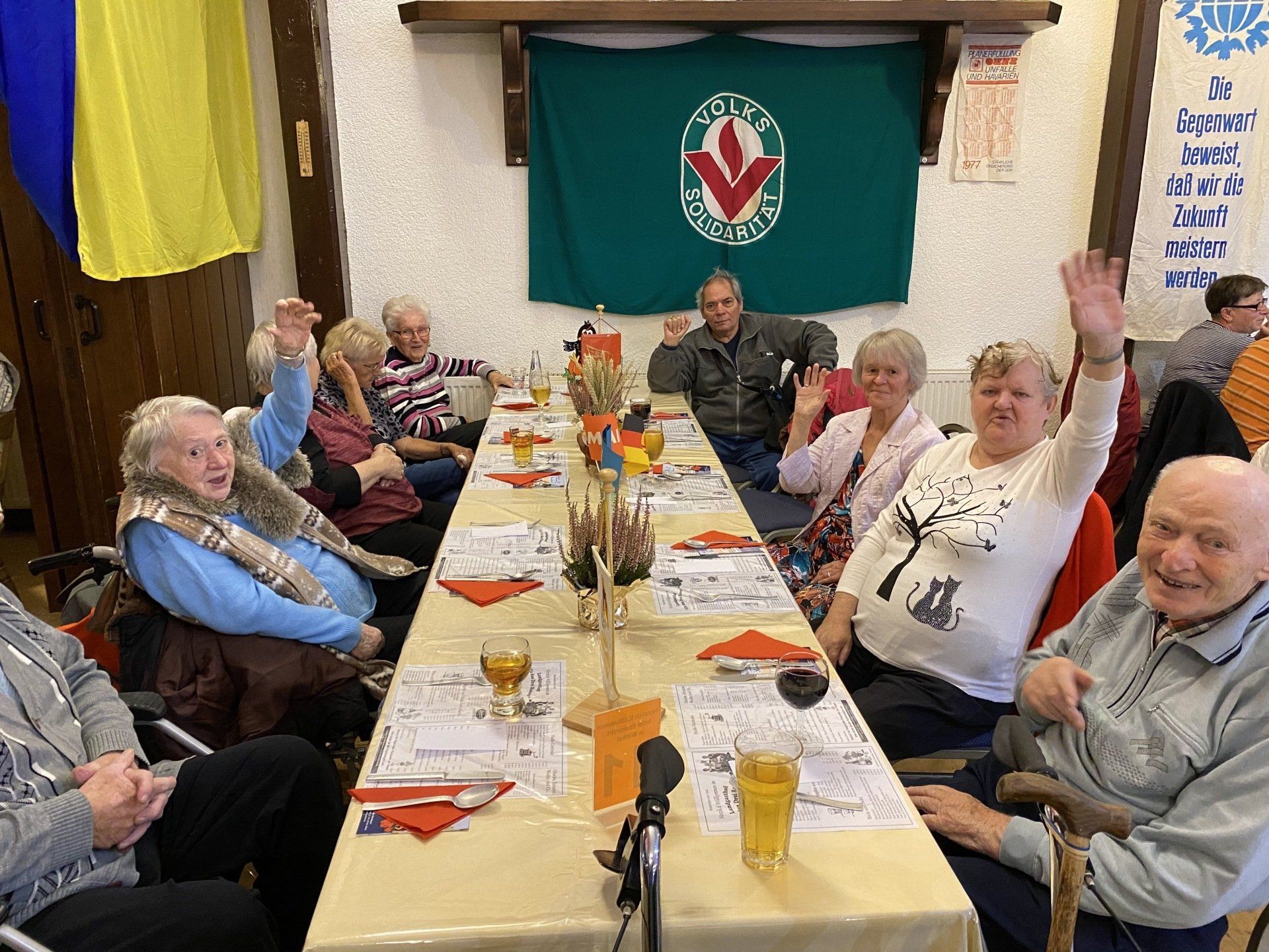 Menschen sitzen an einem langen Tisch, manche winken. Im Hintergrund hängt ein grünes Banner mit einem Logo.