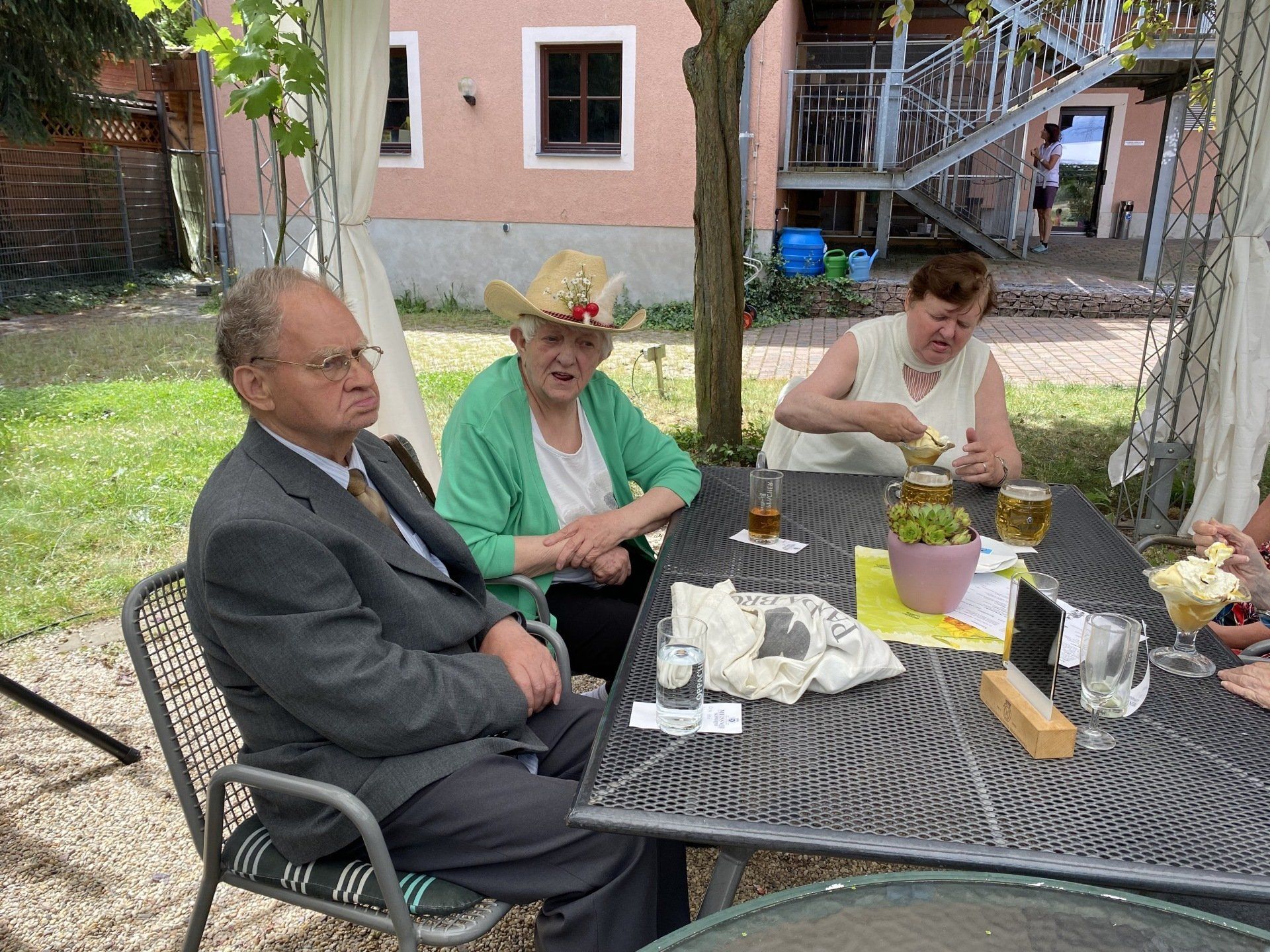 Drei Personen sitzen an einem Tisch im Freien. Einer trägt einen Anzug, ein anderer einen Cowboyhut und der dritte isst.