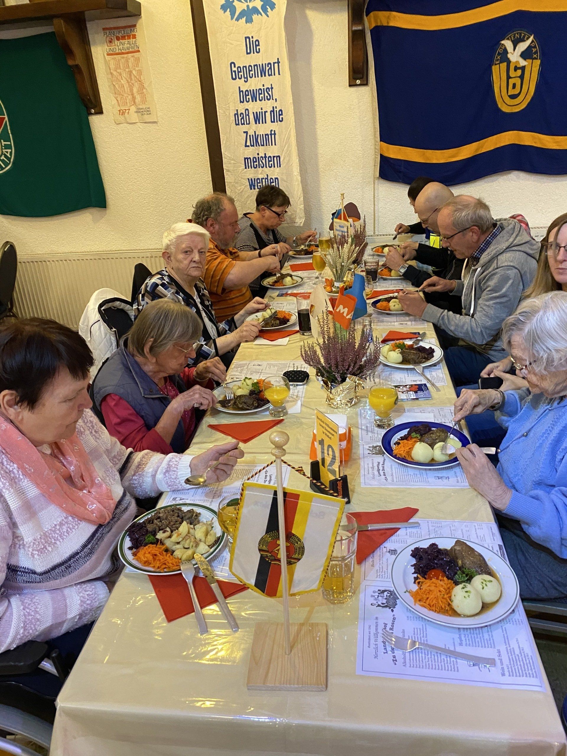 Menschen sitzen an einem langen Tisch und essen. Hinter ihnen hängen Fahnen. 