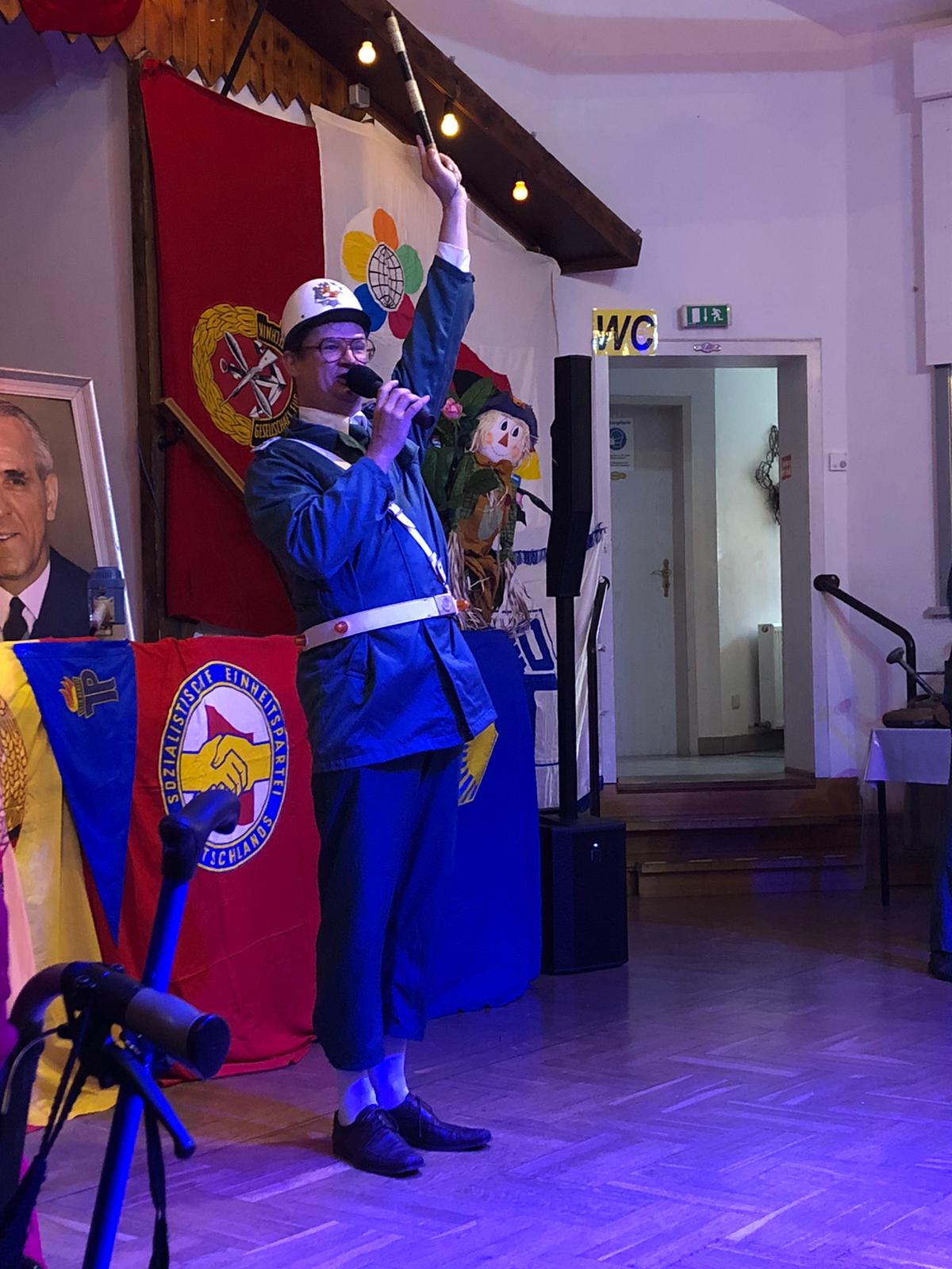 Mann in blauer Uniform mit Mikrofon und Zauberstab tritt auf der Bühne auf. Flaggen und Porträt im Hintergrund.