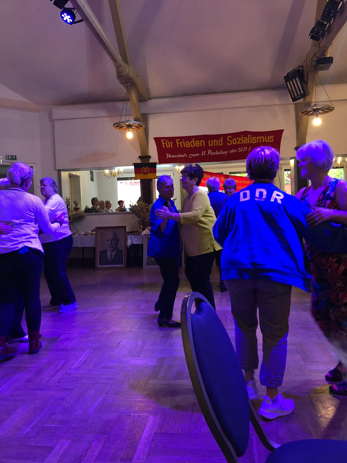 Menschen tanzen in einem Saal. Eine Person trägt eine Jacke mit der Aufschrift „DDR“ auf dem Rücken. 