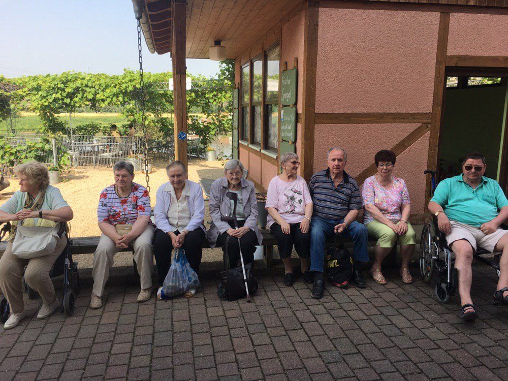 Eine Gruppe von Menschen sitzt auf einer Bank im Freien. Einige benutzen Rollstühle. Gebäude mit Holzbalken und Ziegelweg.
