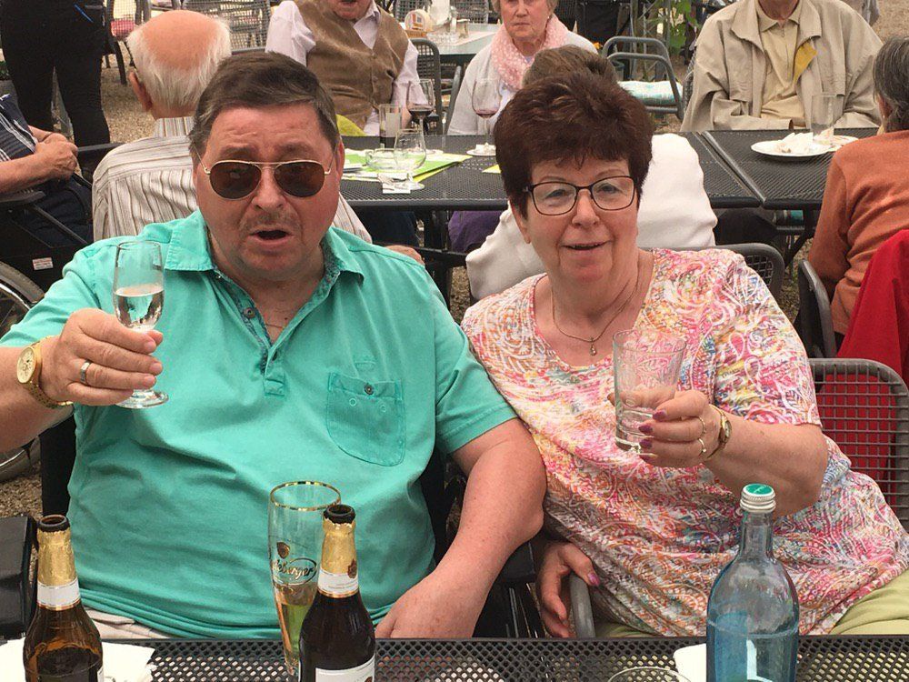 Paar im Freien, stößt mit Getränken an. Mann in grünem Hemd, Sonnenbrille; Frau in geblümtem Top.