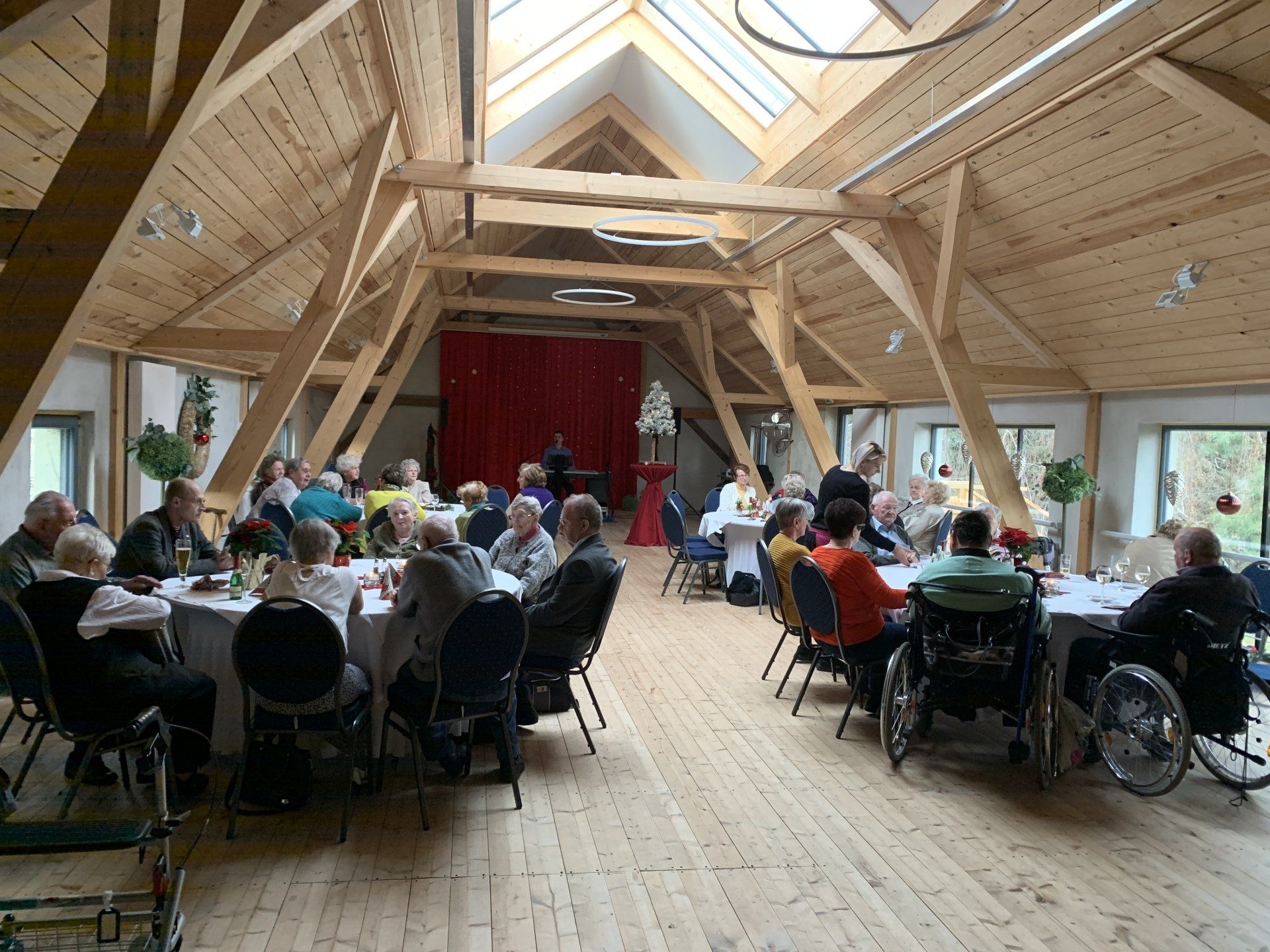 Menschen sitzen an Tischen in einem großen Raum mit freiliegenden Holzbalken, im Hintergrund eine Bühne.
