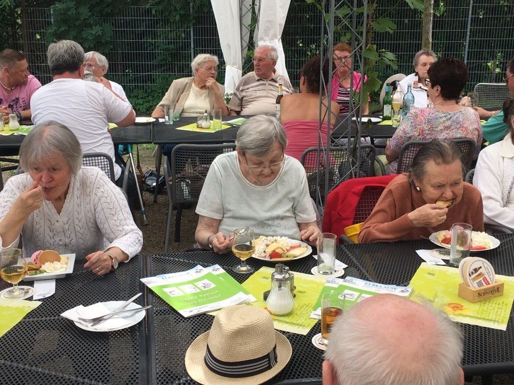 Menschen essen und trinken an Tischen im Freien unter einem Baldachin. Einige lächeln und eine Person trägt einen Hut.