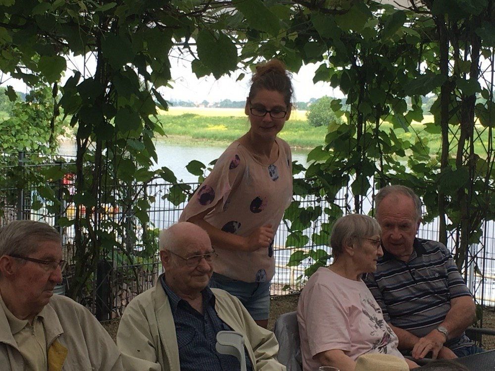 Menschen sitzen an einem Tisch unter Weinreben in der Nähe eines Gewässers. Hinter ihnen steht eine lächelnde Frau.