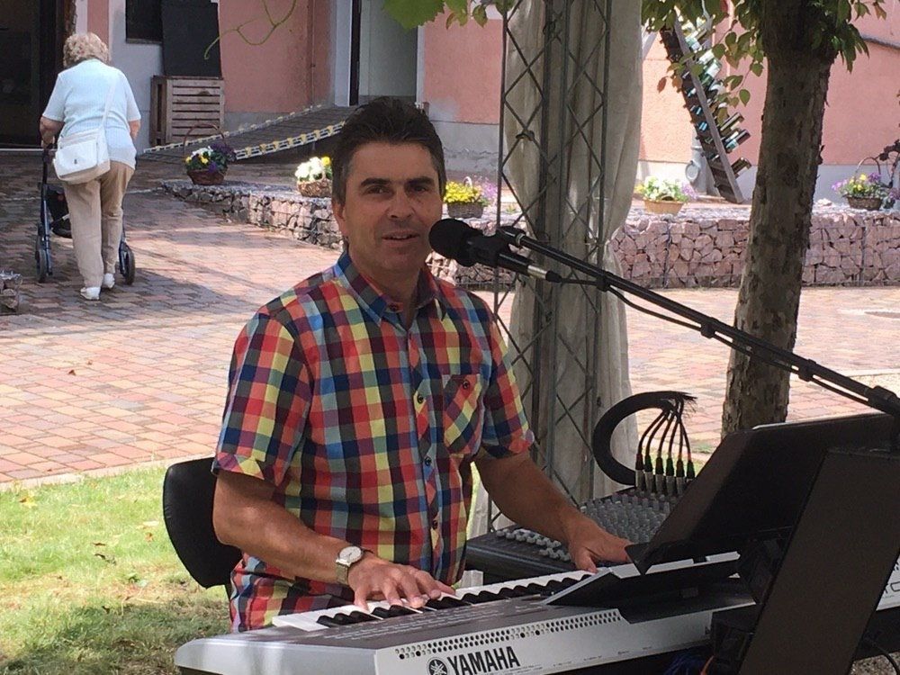 Mann spielt Keyboard und singt draußen; ältere Frau geht im Hintergrund.