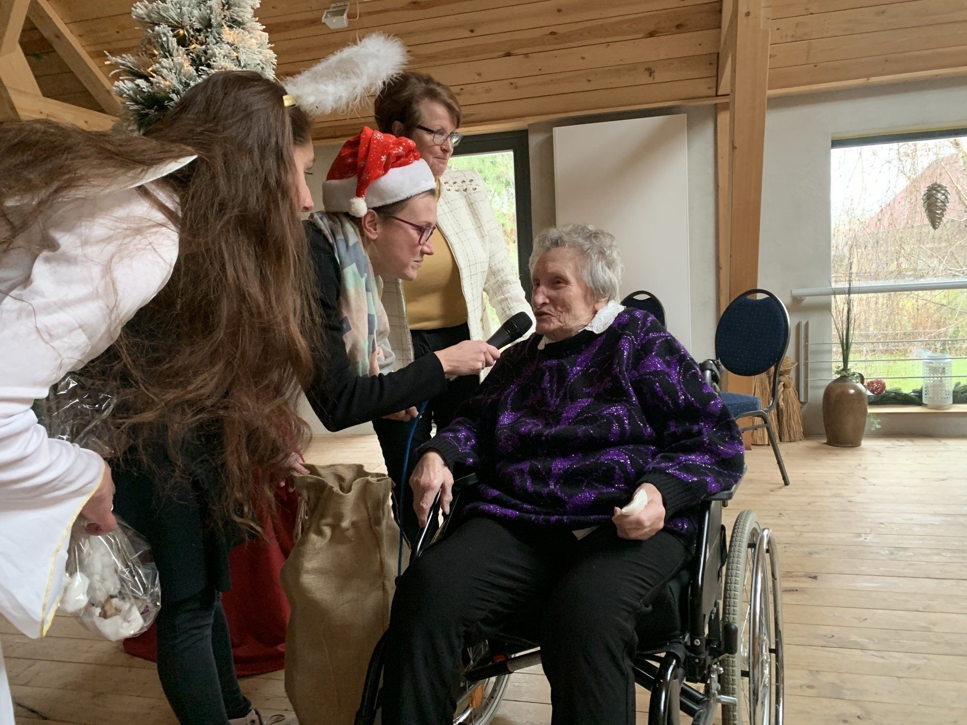 Frau im Rollstuhl wird drinnen von Menschen mit Engelsflügeln und Weihnachtsmannmütze interviewt.