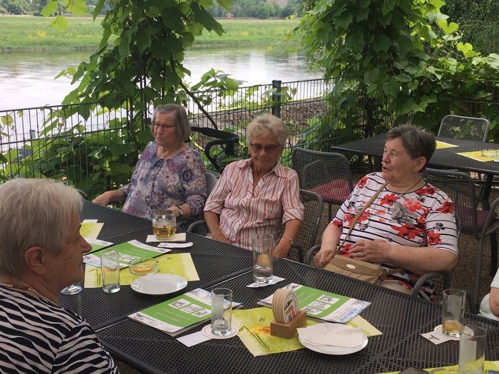 Eine Gruppe von Frauen sitzt an einem Tisch am Fluss und genießt Getränke und Gespräche im Freien.