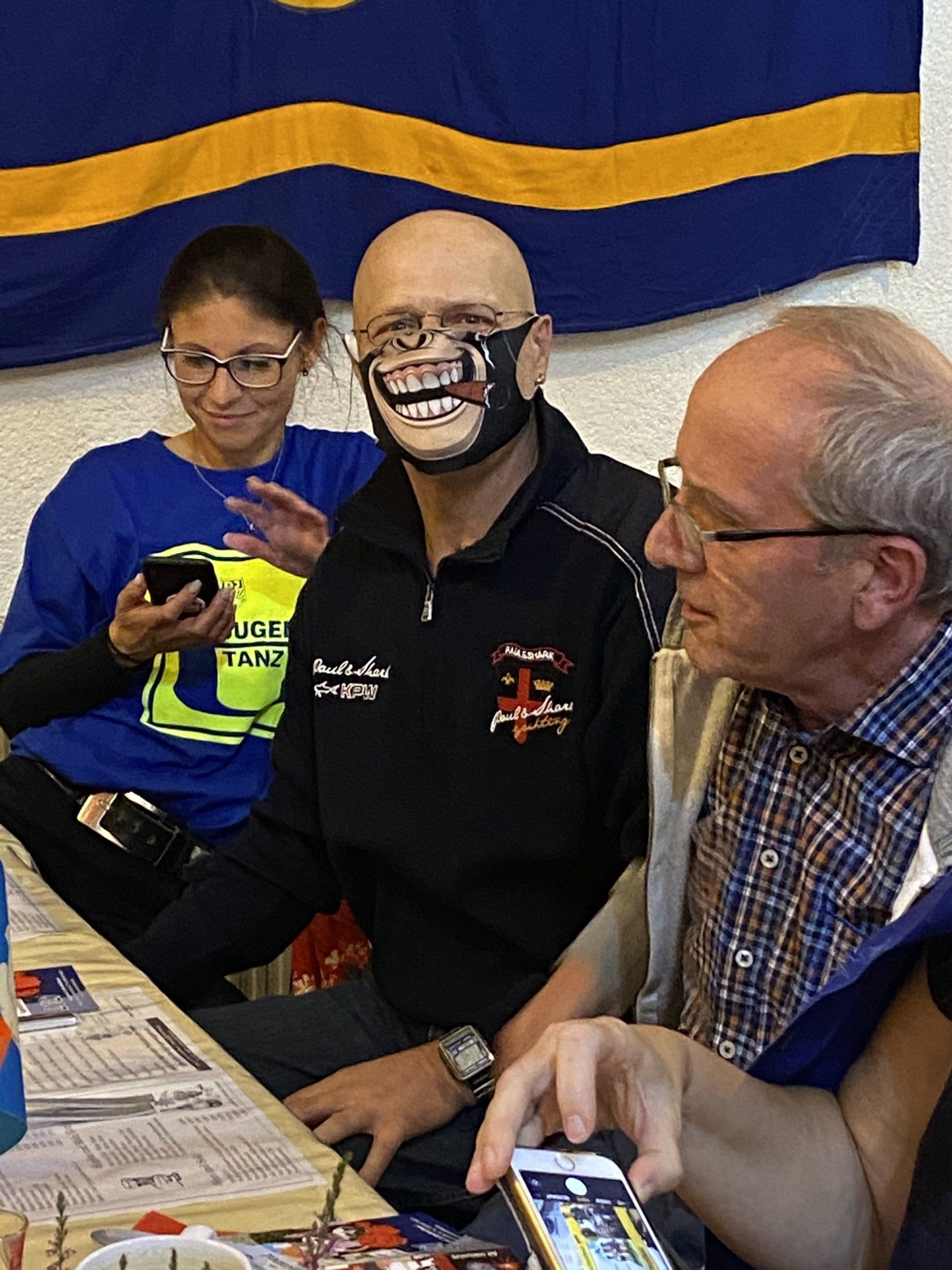 Drei Personen sitzen an einem Tisch. In der Mitte trägt ein Mann eine Gesichtsmaske mit einem lächelnden Mund. 