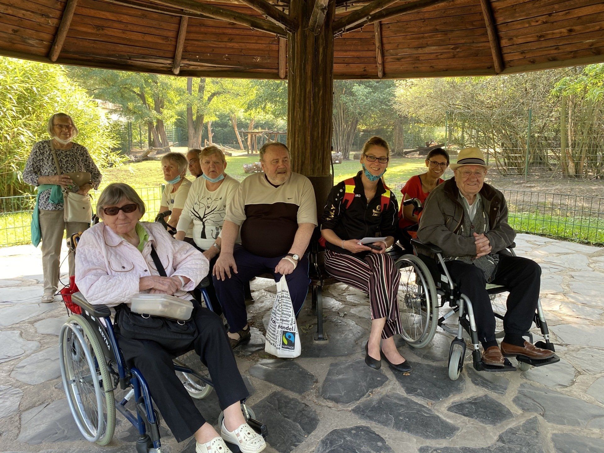 Eine Gruppe von Menschen sitzt in einem Pavillon, einige davon im Rollstuhl. Sie scheinen sich in einem Park auszuruhen.