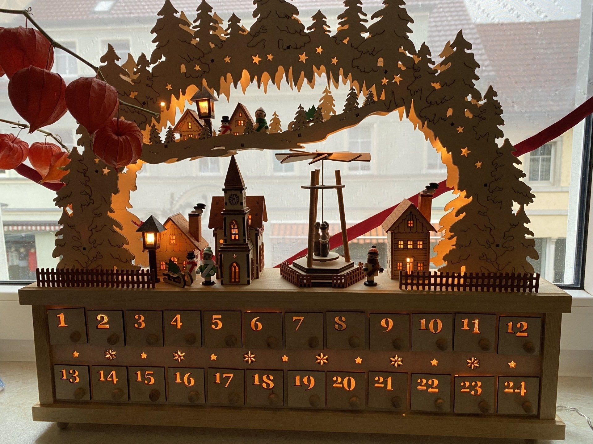 Adventskalender aus Holz mit beleuchteten Häusern, Bäumen und Zahlen.