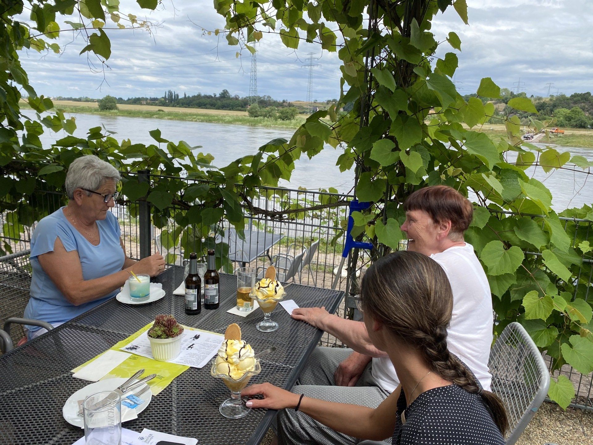 Drei Personen sitzen an einem Tisch und essen Eis mit Blick auf den Fluss. Weinblätter über ihnen.