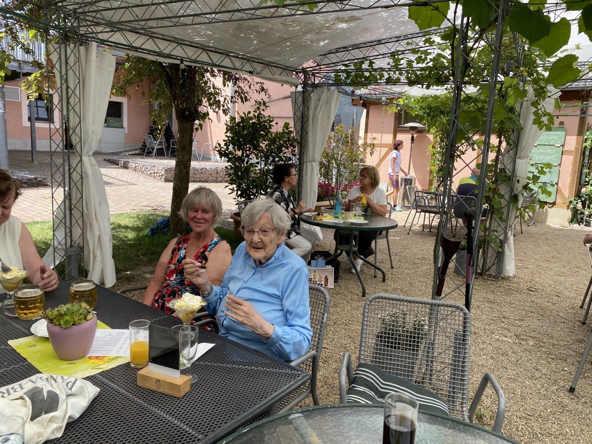 Menschen sitzen an Tischen im Freien unter einer Pergola und genießen Getränke und Essen. Gartenkulisse.