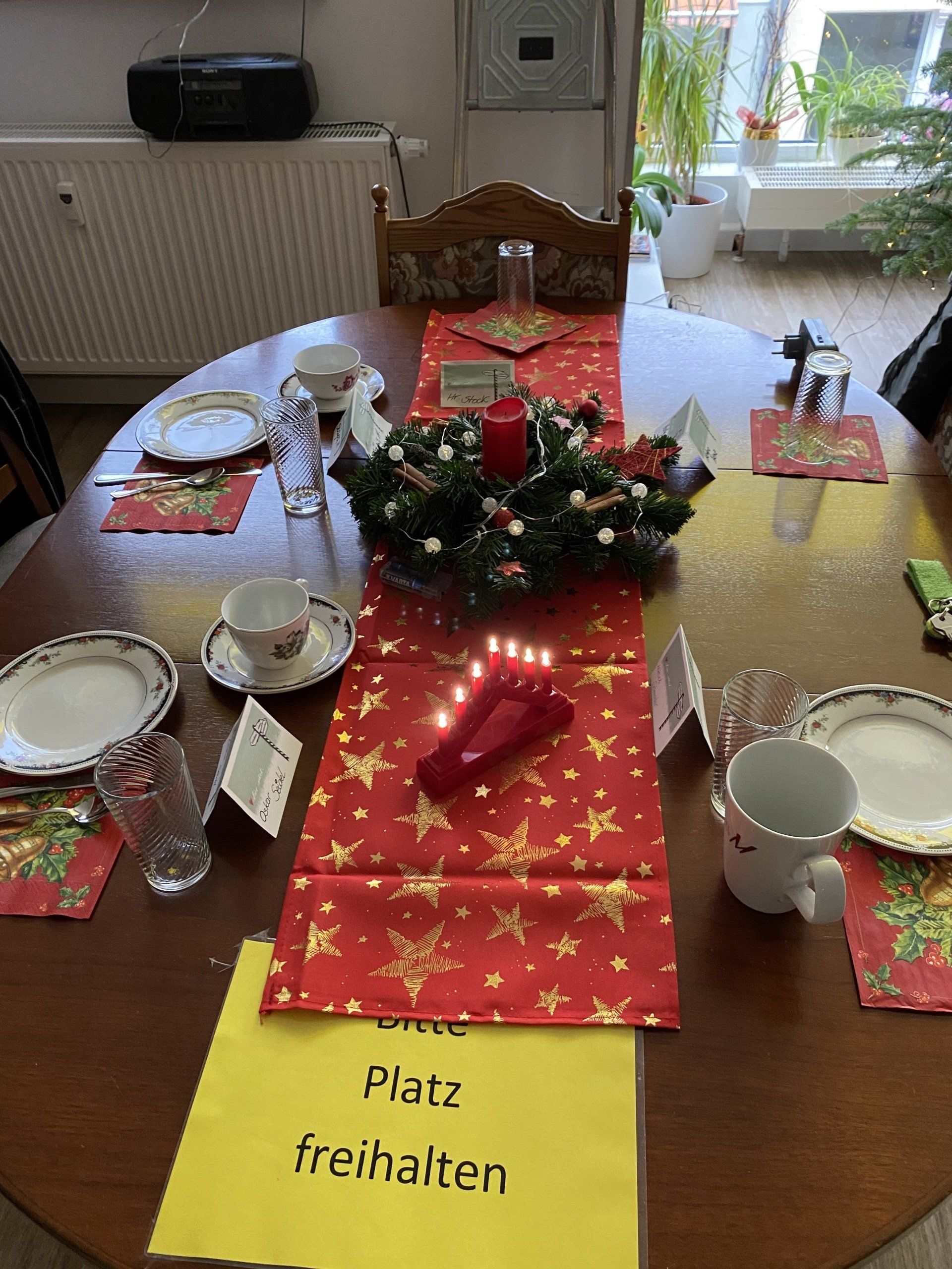 Festlich gedeckter Tisch mit rotem Tischläufer, Kerzenkranz und Gedecken. Schild „Platz freihalten“.