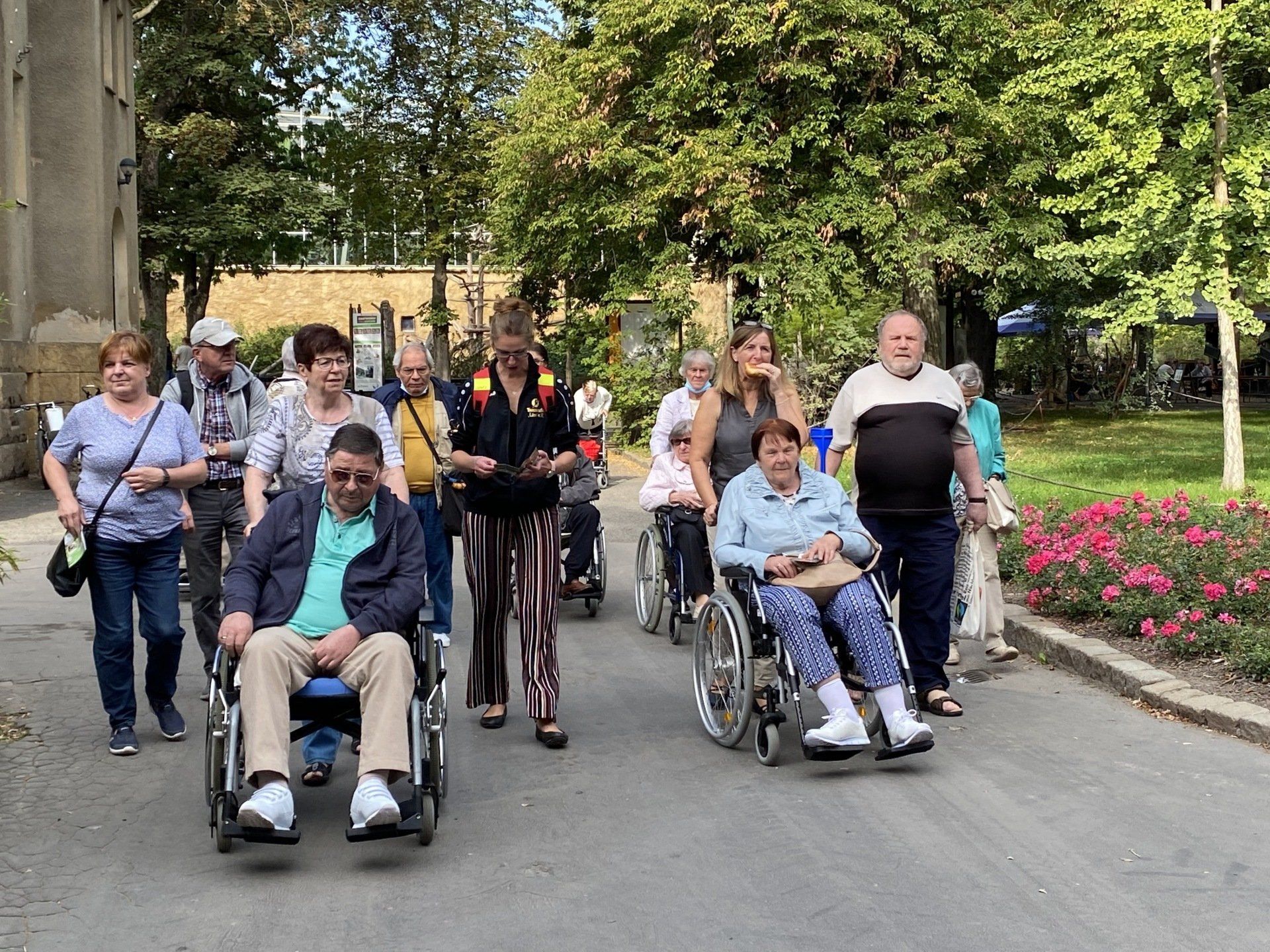 Eine Gruppe von Menschen, einige davon im Rollstuhl, geht einen von Bäumen und einem Gebäude gesäumten Weg entlang.