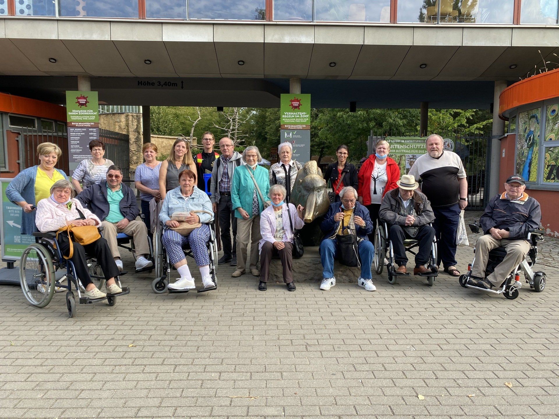 Eine Gruppe von Menschen, einige davon im Rollstuhl, posiert vor einem Gebäude mit grünen Schildern und einem Betonweg.