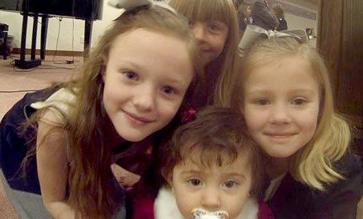 Four smiling children huddle closely together indoors, with a toddler in front holding a pacifier.