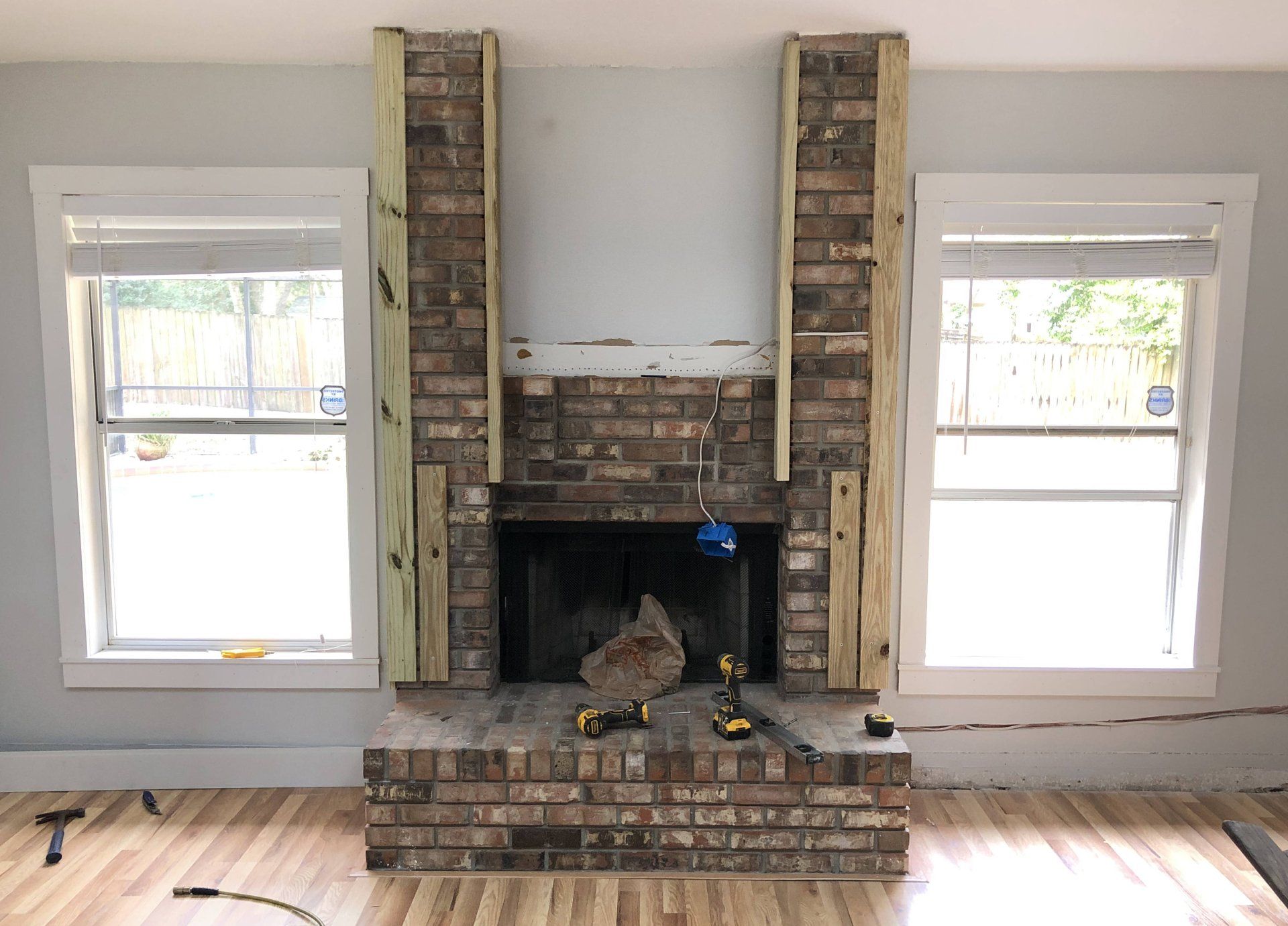 Brick fireplace with wood trim being added, flanked by windows.