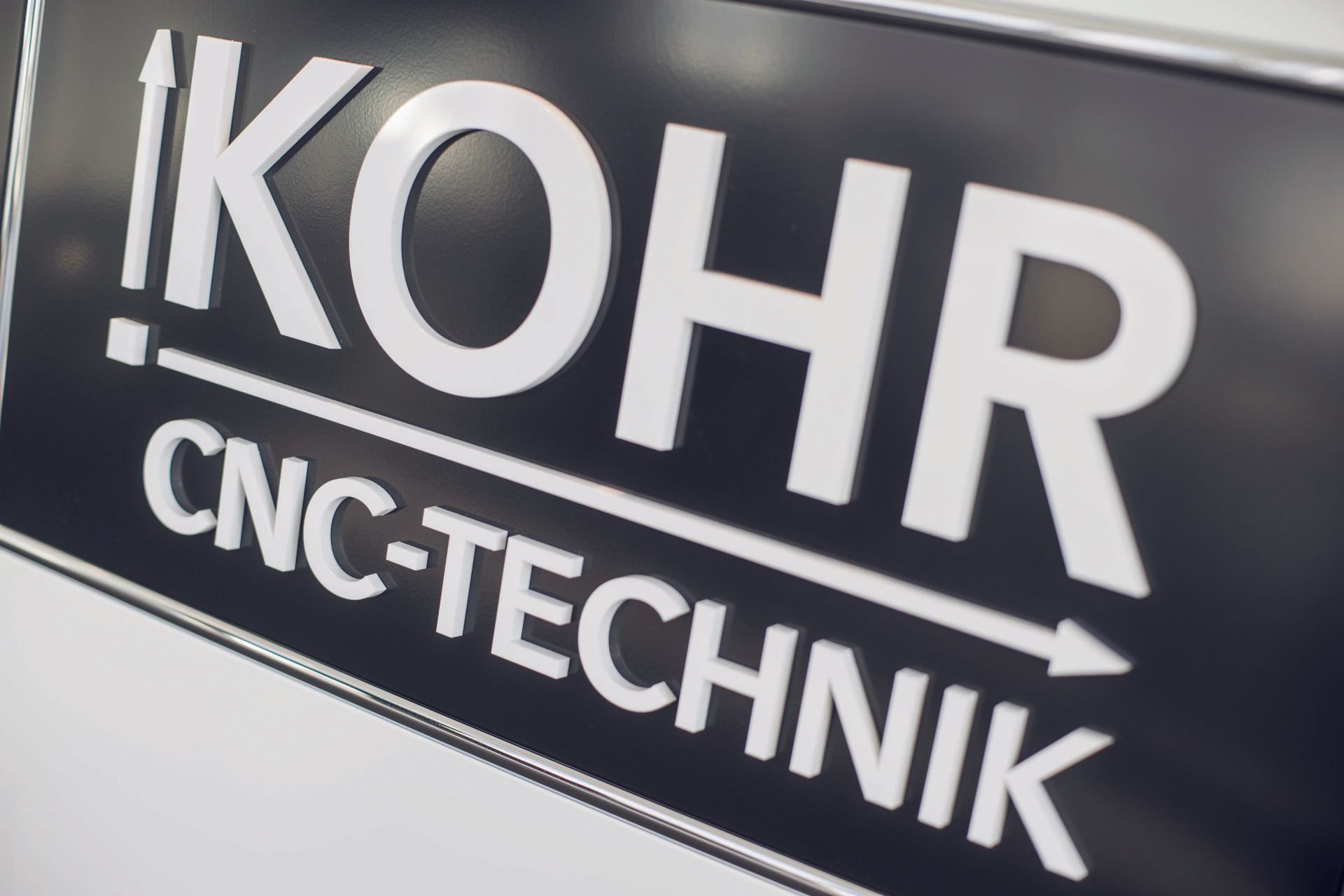 Schwarzes Schild mit weißer Schrift und der Aufschrift „KOHR CNC-TECHNIK“. Pfeilgrafik neben „KOHR“.
