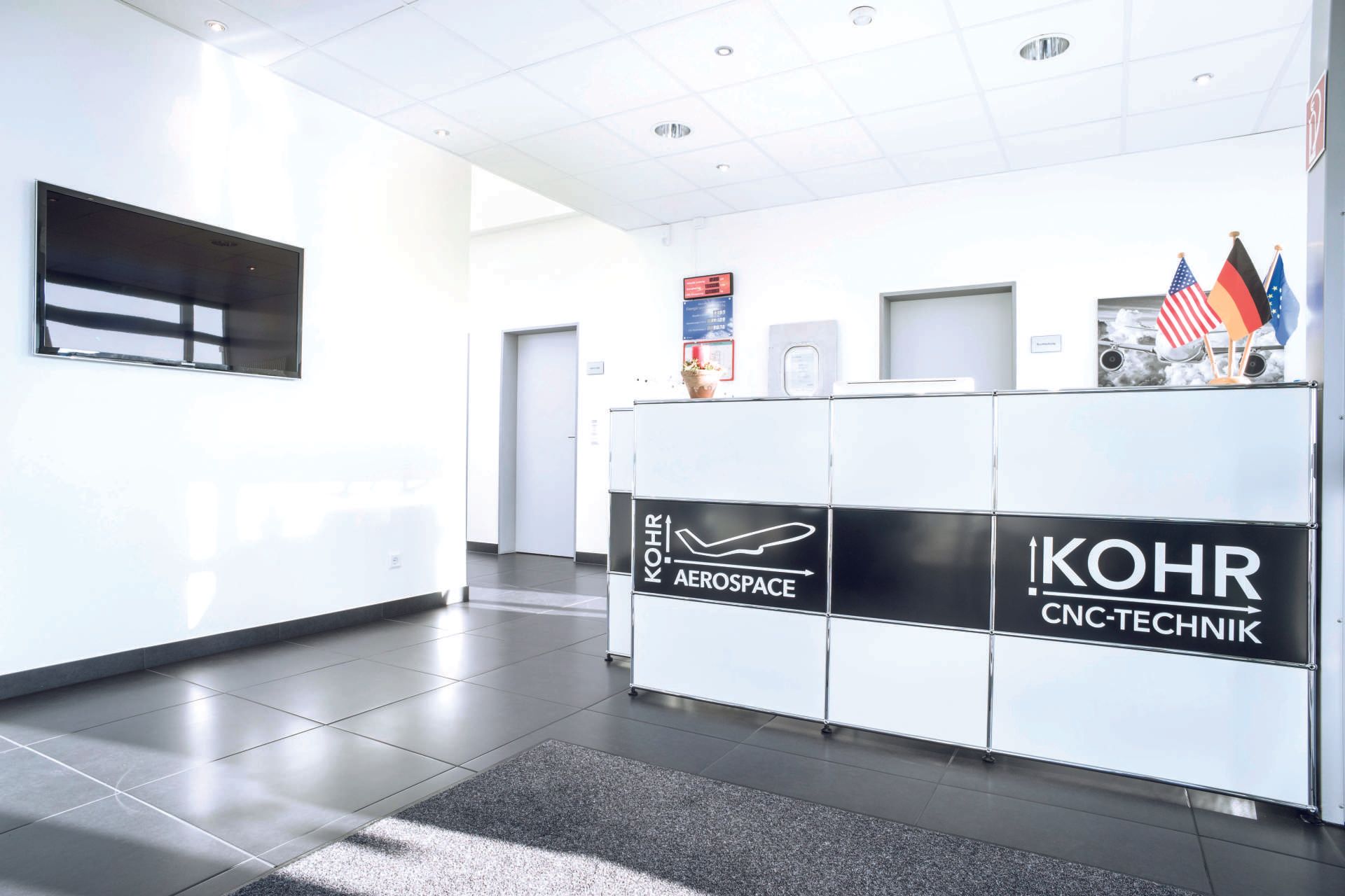 Empfangsbereich mit weißem Schreibtisch und Logo von IKOHR CNC-Technik, mit Fahnen und einem Fernseher an der Wand.