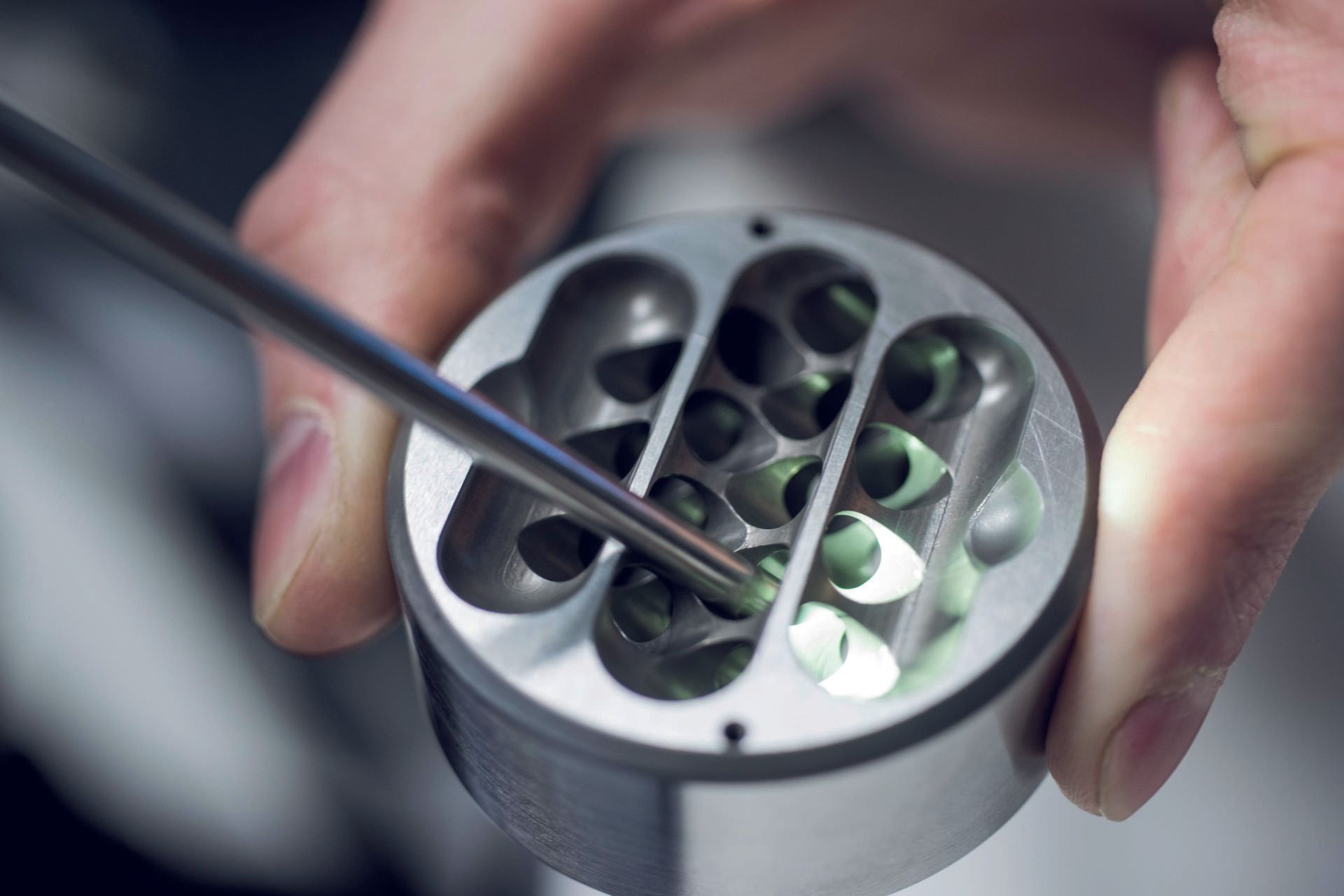 Nahaufnahme einer Hand, die mit einem Werkzeug kleine, grün leuchtende Objekte in einem Metallzylinder manipuliert.