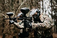 paintball sport player wearing protective mask aiming gun from shelter under gunfire attack with paint splash