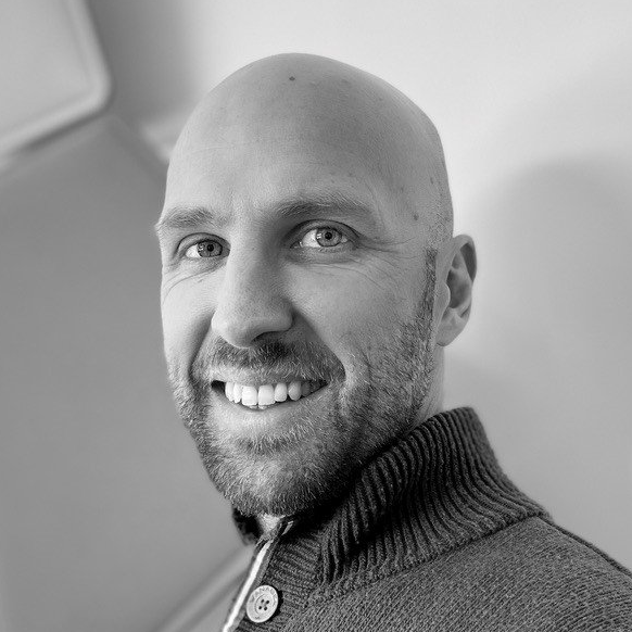 Un homme chauve souriant, vêtu d'un pull foncé, pose à l'intérieur devant un mur clair.