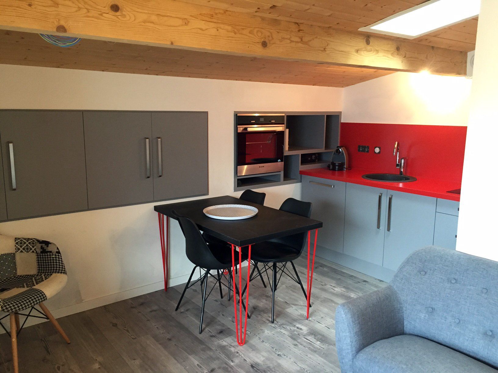 Cuisine et salle à manger modernes avec placards gris, crédence rouge, table noire et plafond en bois apparent.