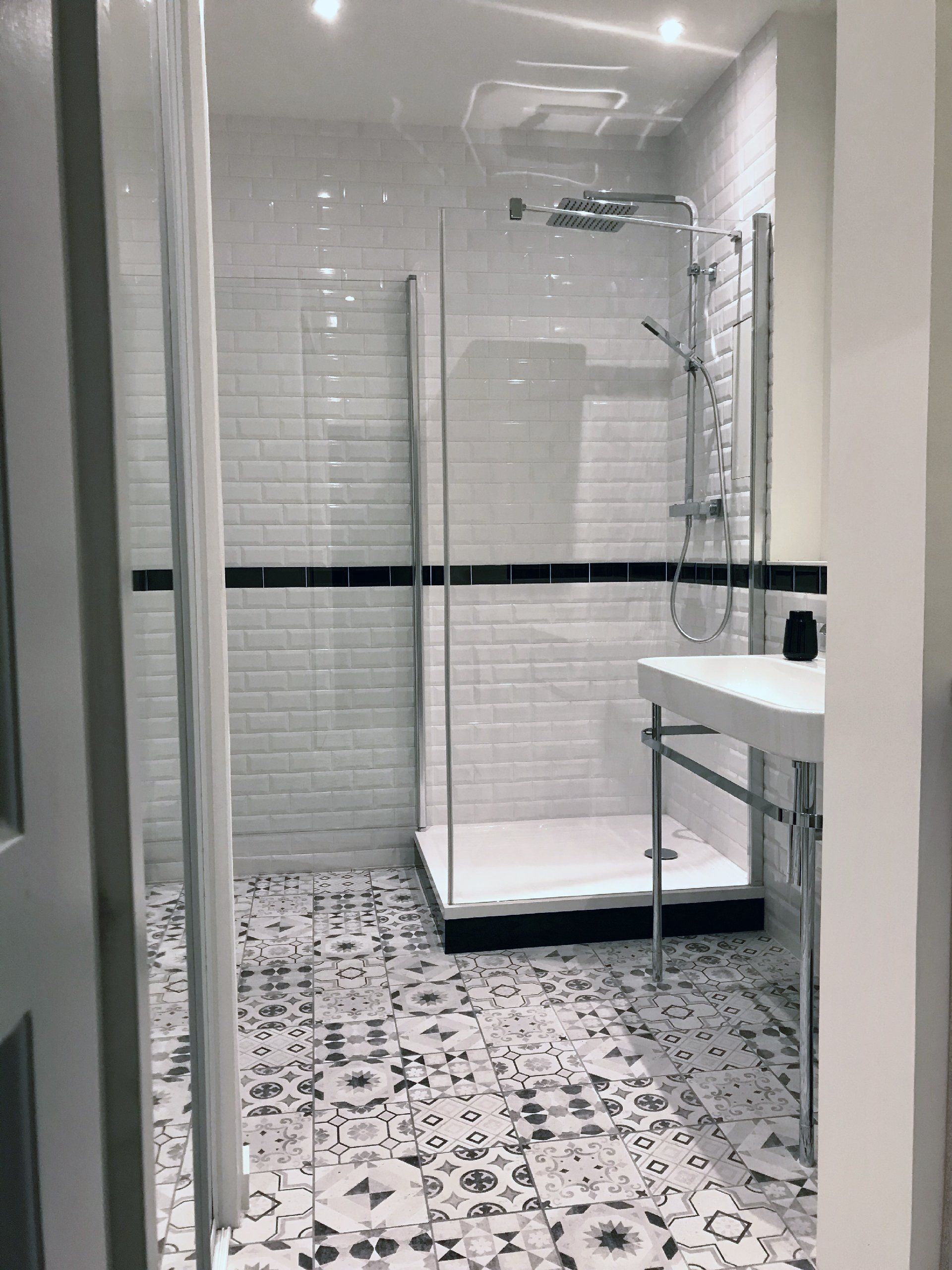 Salle de bains moderne avec douche en verre, lavabo blanc, carrelage noir décoratif et sol à motifs.