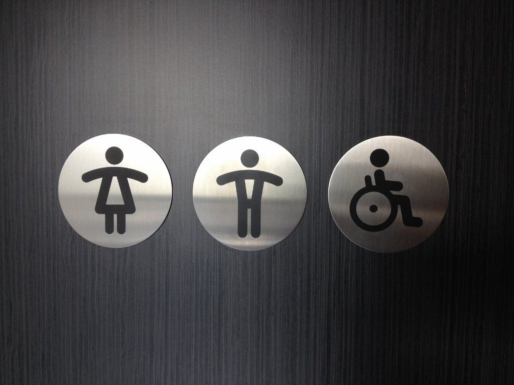 Trois panneaux argentés pour les toilettes femmes, hommes et accès pour fauteuils roulants, sur un mur sombre.