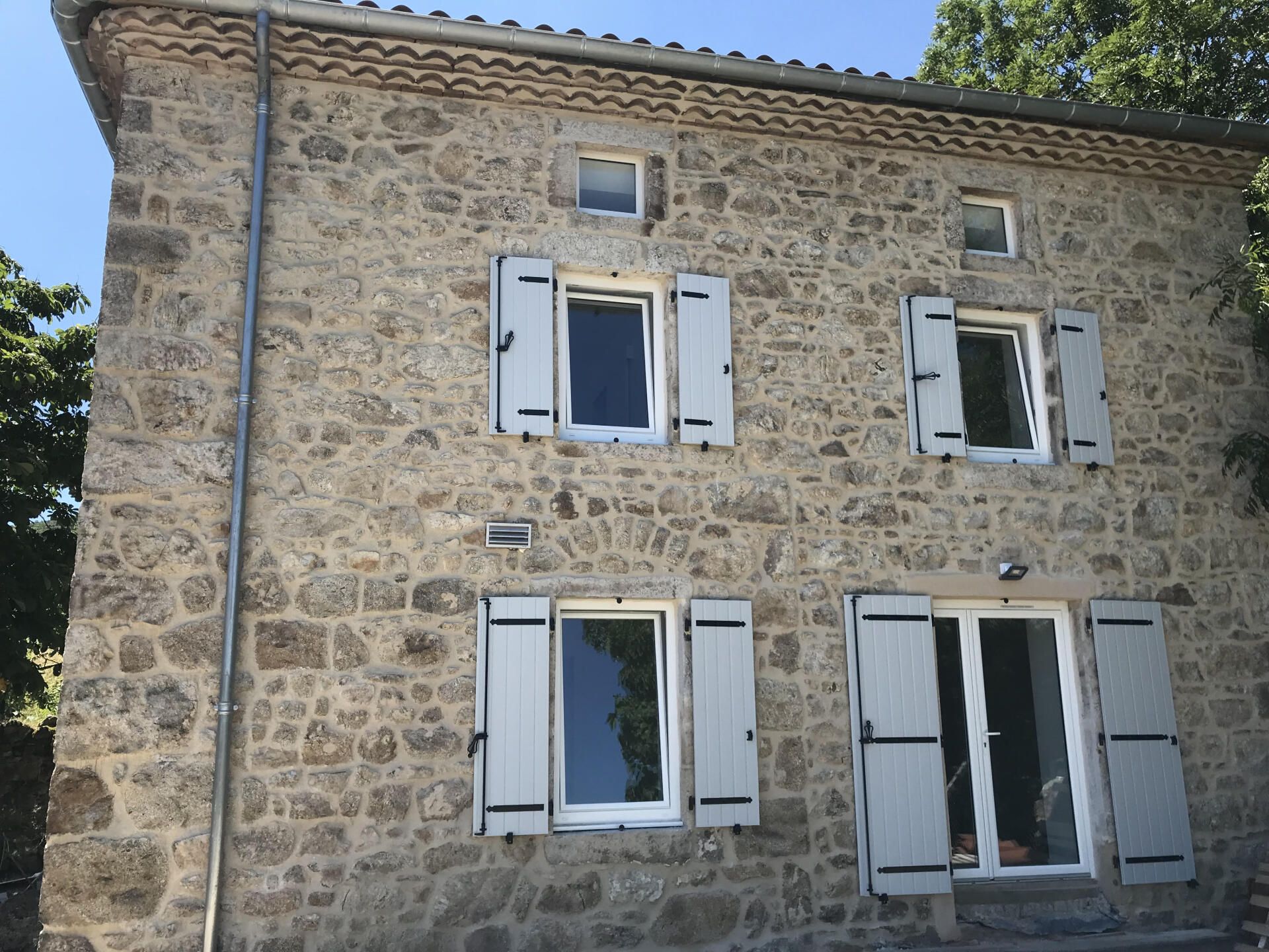 Façade de maison en pierre avec volets blancs et quatre fenêtres sous un toit de tuiles