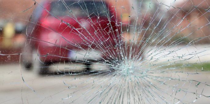A close up of a broken windshield with a red car in the background.