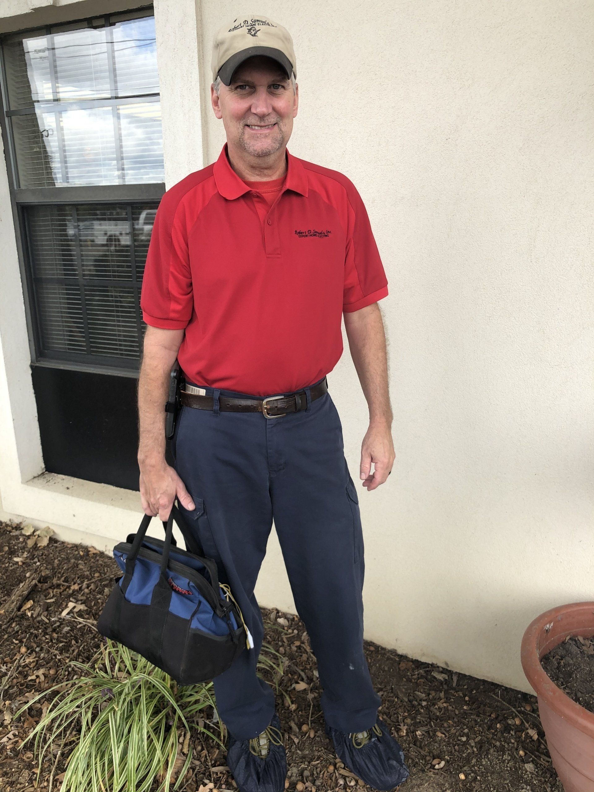 Man in red shirt and blue pants, holding a tool bag, standing outside a building.