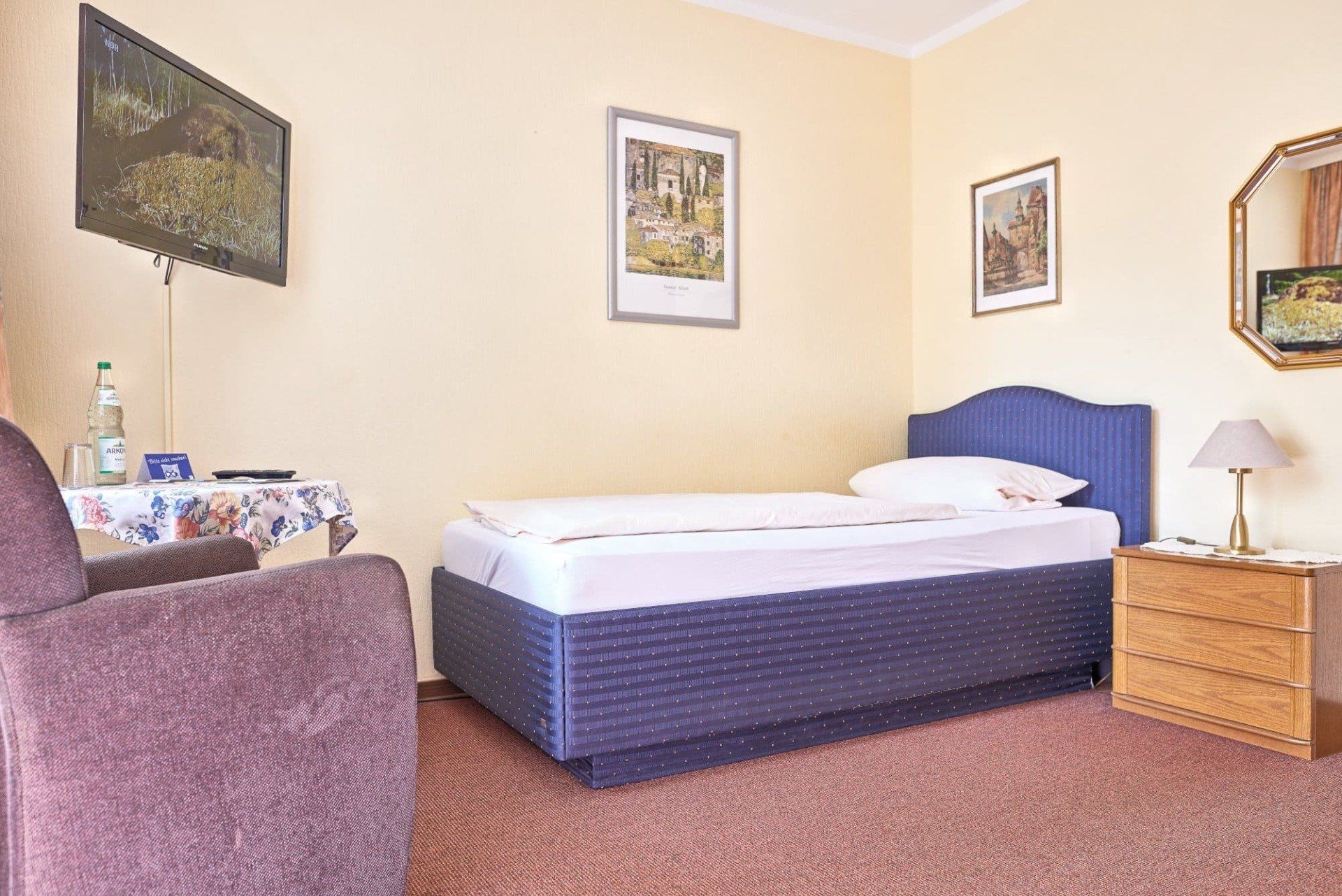 Ein Hotelzimmer mit Einzelbett, Sessel, Nachttisch und Fernseher.