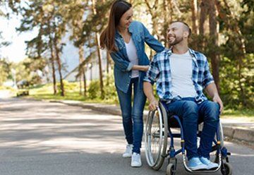 Woman pushes smiling man in a wheelchair along a sunlit park path — Health Experts In Toowoomba, QLD