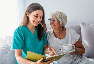Two women on a couch, smiling over a book and cup of tea in a bright living room — Health Experts In Toowoomba, QLD