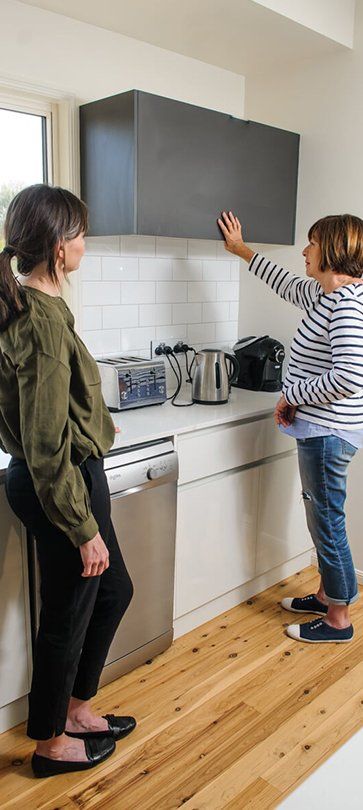 Two people in a small kitchen adjusting a dark wall cabinet near a gas stove — Health Experts In Toowoomba, QLD