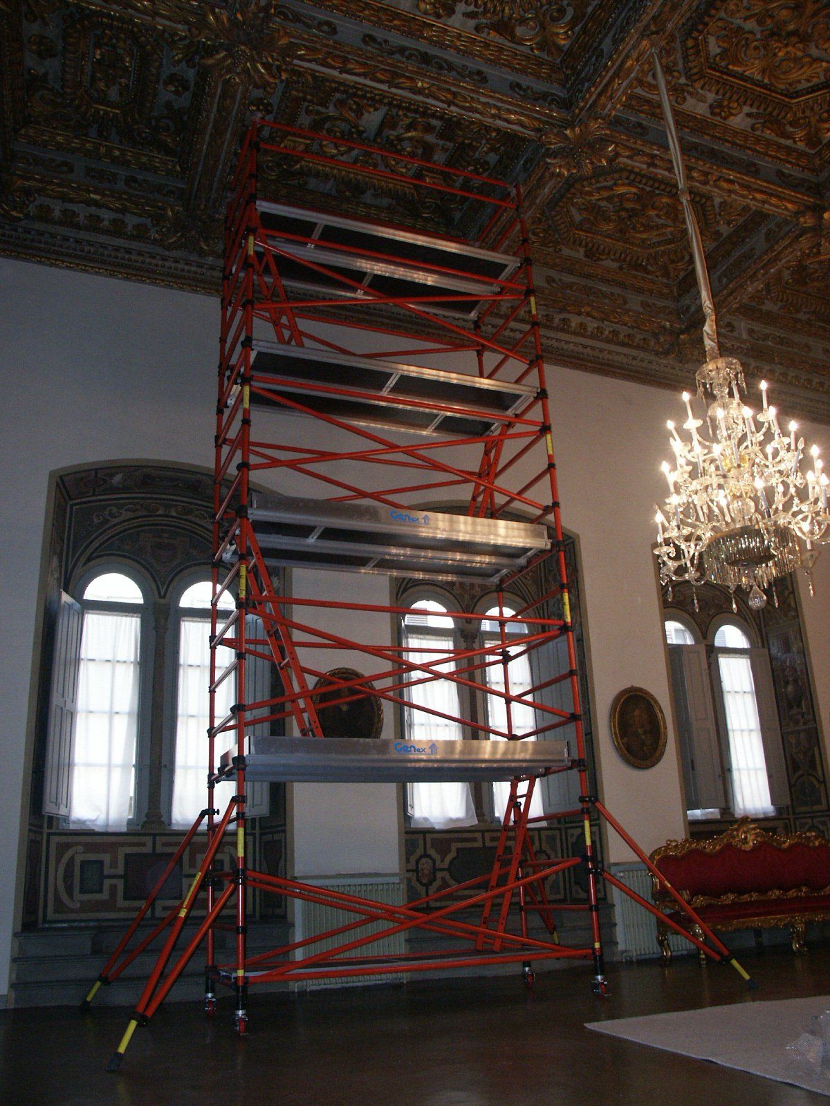 A red scaffolding is in a large room with a chandelier hanging from the ceiling.