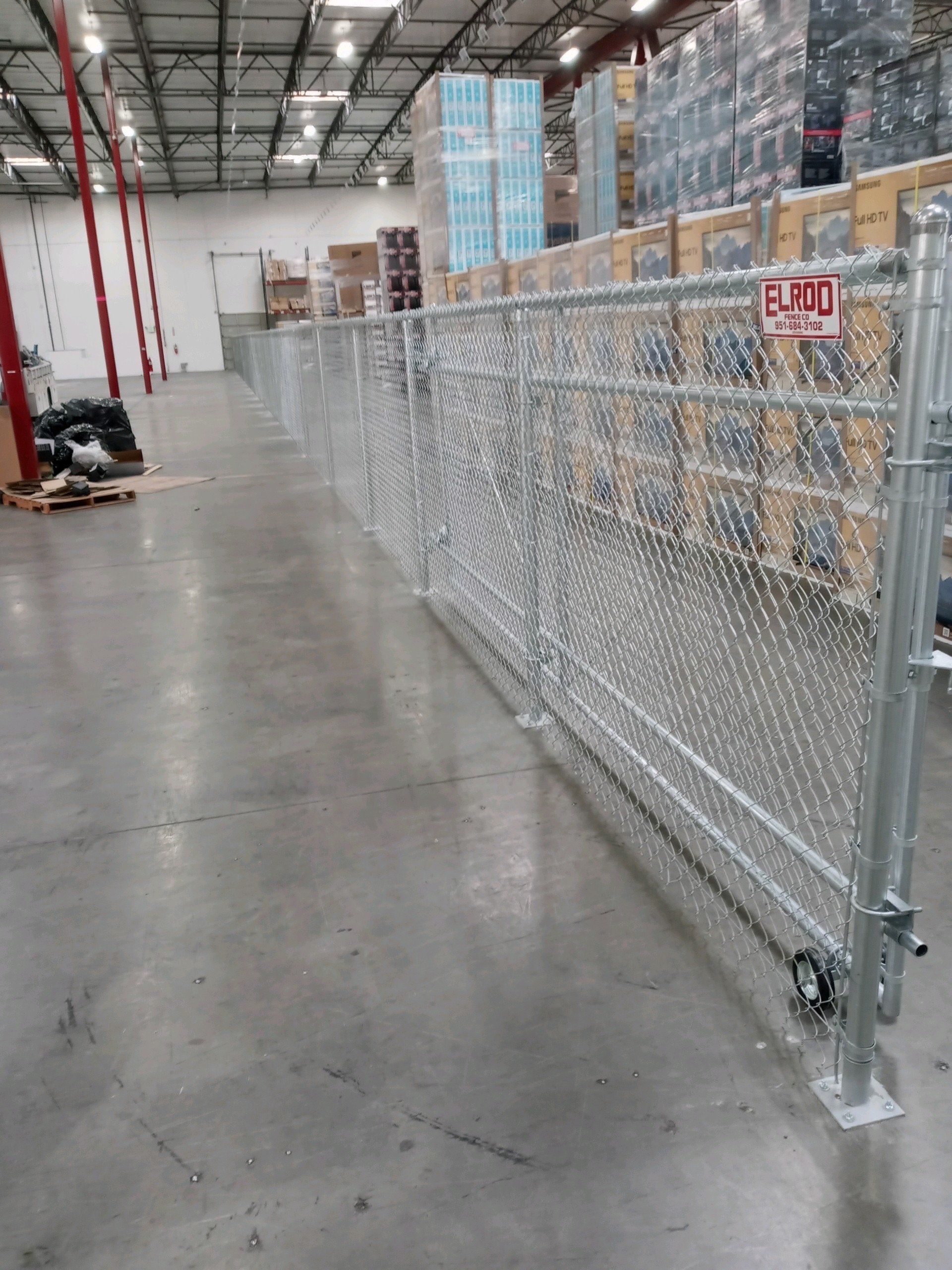 Chain-link fence inside a warehouse, separating aisles. Pallets and boxes in the background.