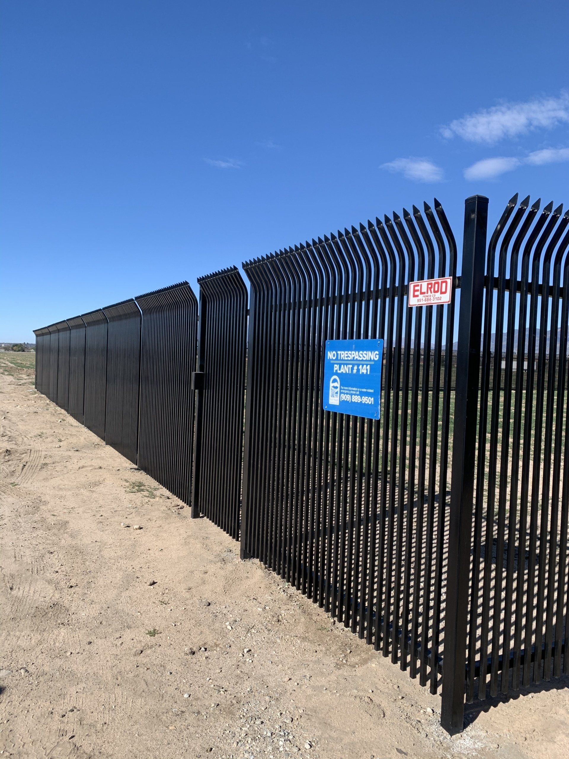 Black security fence with curved vertical bars topped with spikes, blue warning sign. Outdoors, under blue sky.