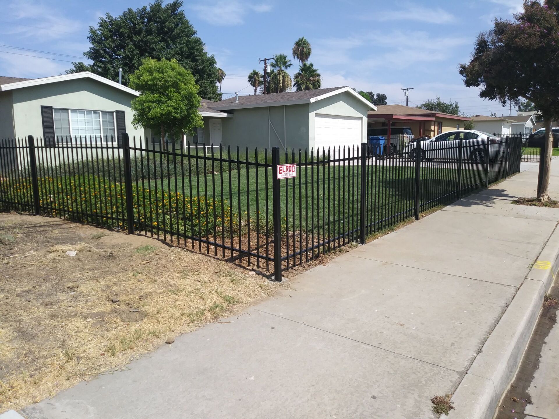 Black metal fence encloses a front yard with green grass and shrubs; houses in the background.
