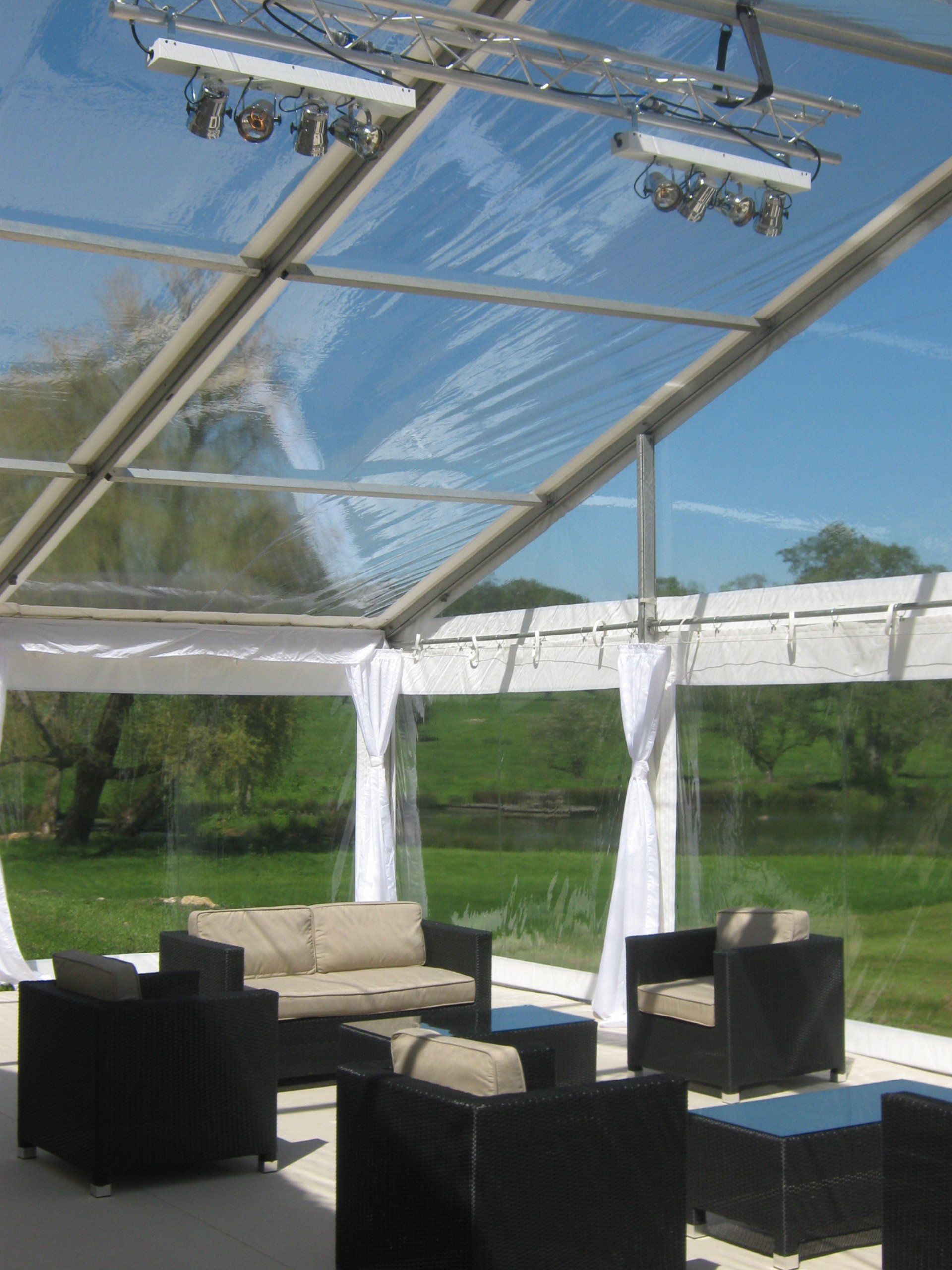 Clear tent interior with seating. Sunlight streams through. White curtains, black armchairs, and beige couch.