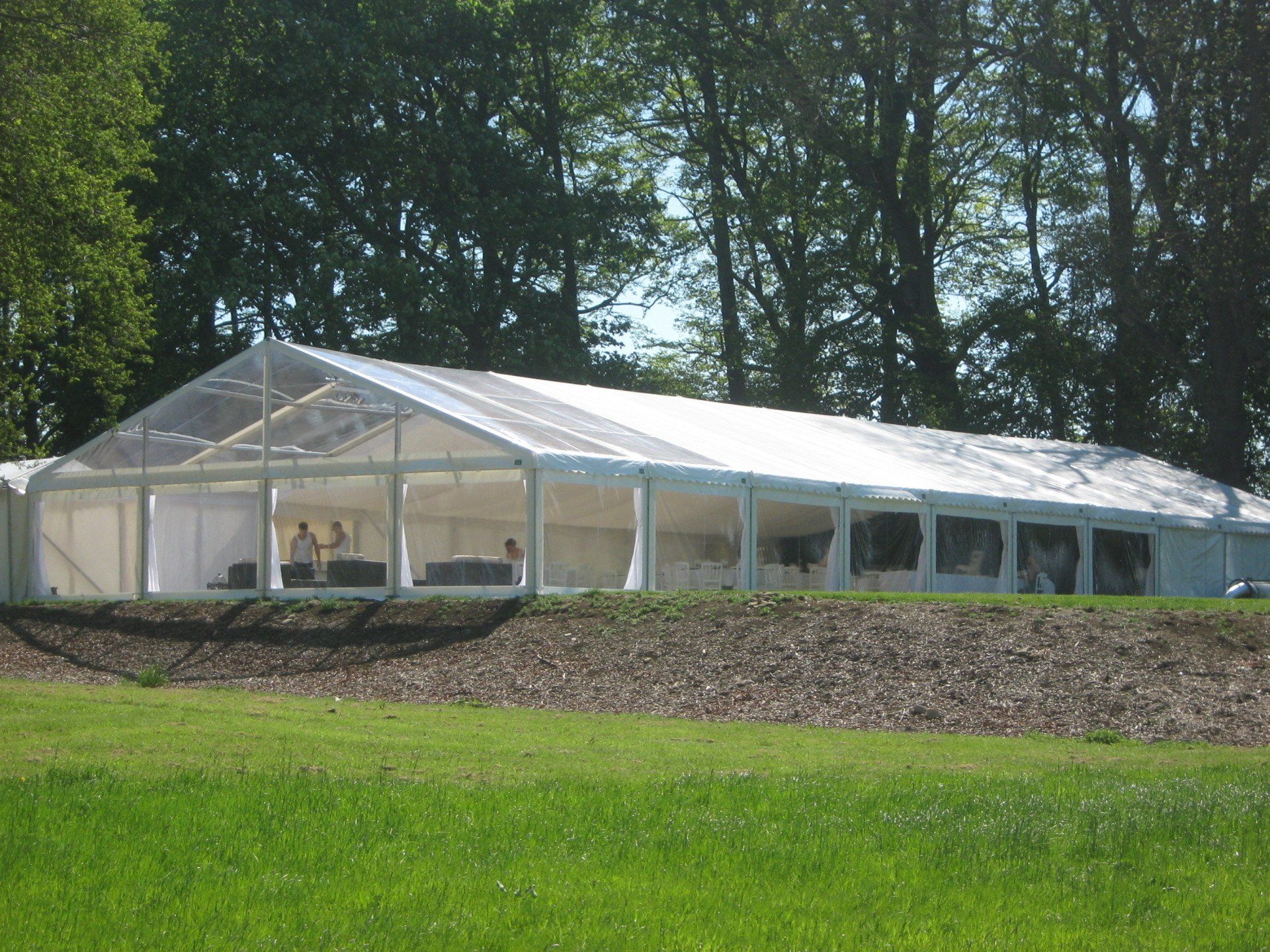 Large clear-roofed tent set up on a grassy field, with people inside. Trees in the background.