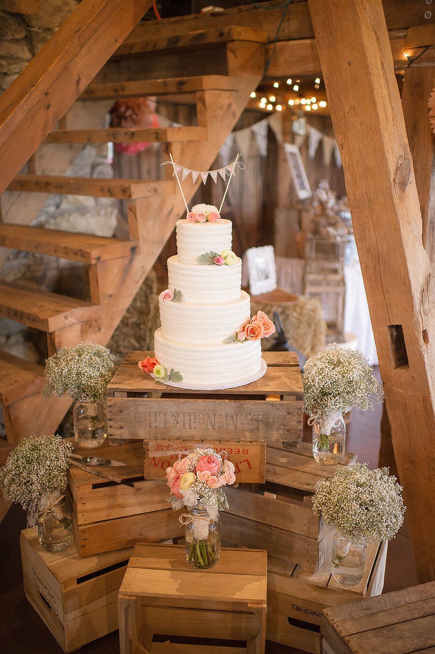 Wedding cake on stacked wooden crates with flower arrangements, under wooden stairs.