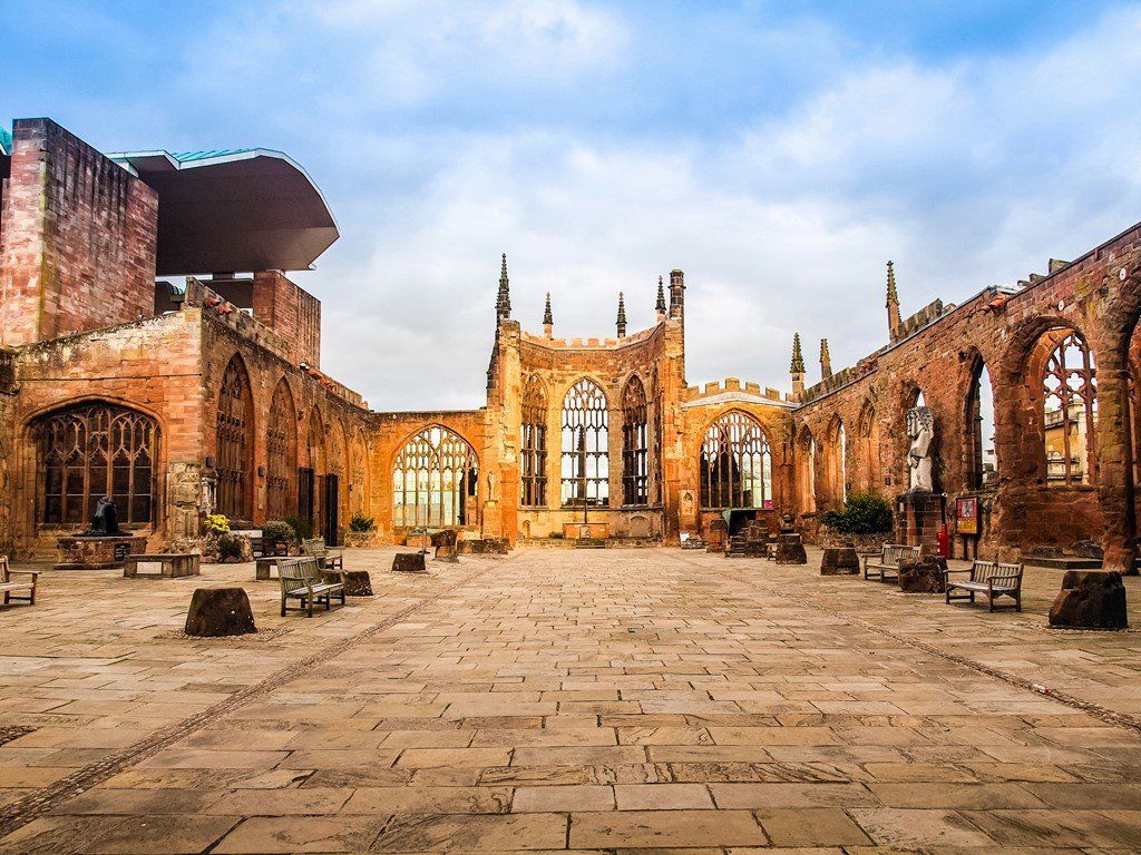 Ruined cathedral exterior in Coventry, England. Brick walls with large arched windows and a paved courtyard. Cloudy sky.
